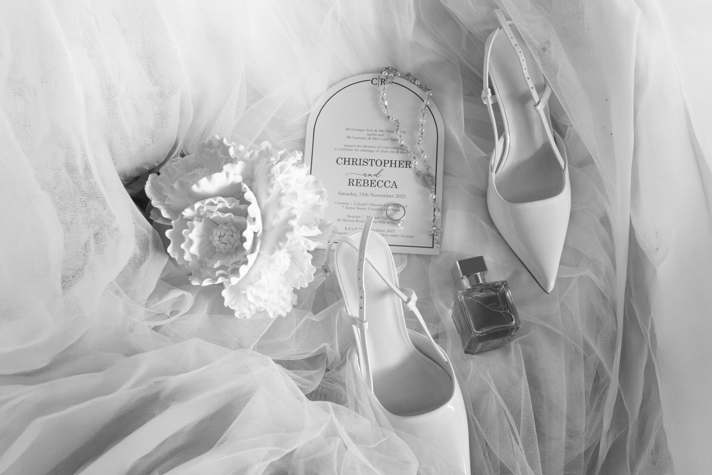 Black and white flat lay of bridal accessories including heels, bouquet, perfume, and a wedding invitation for Christopher and Rebecca.