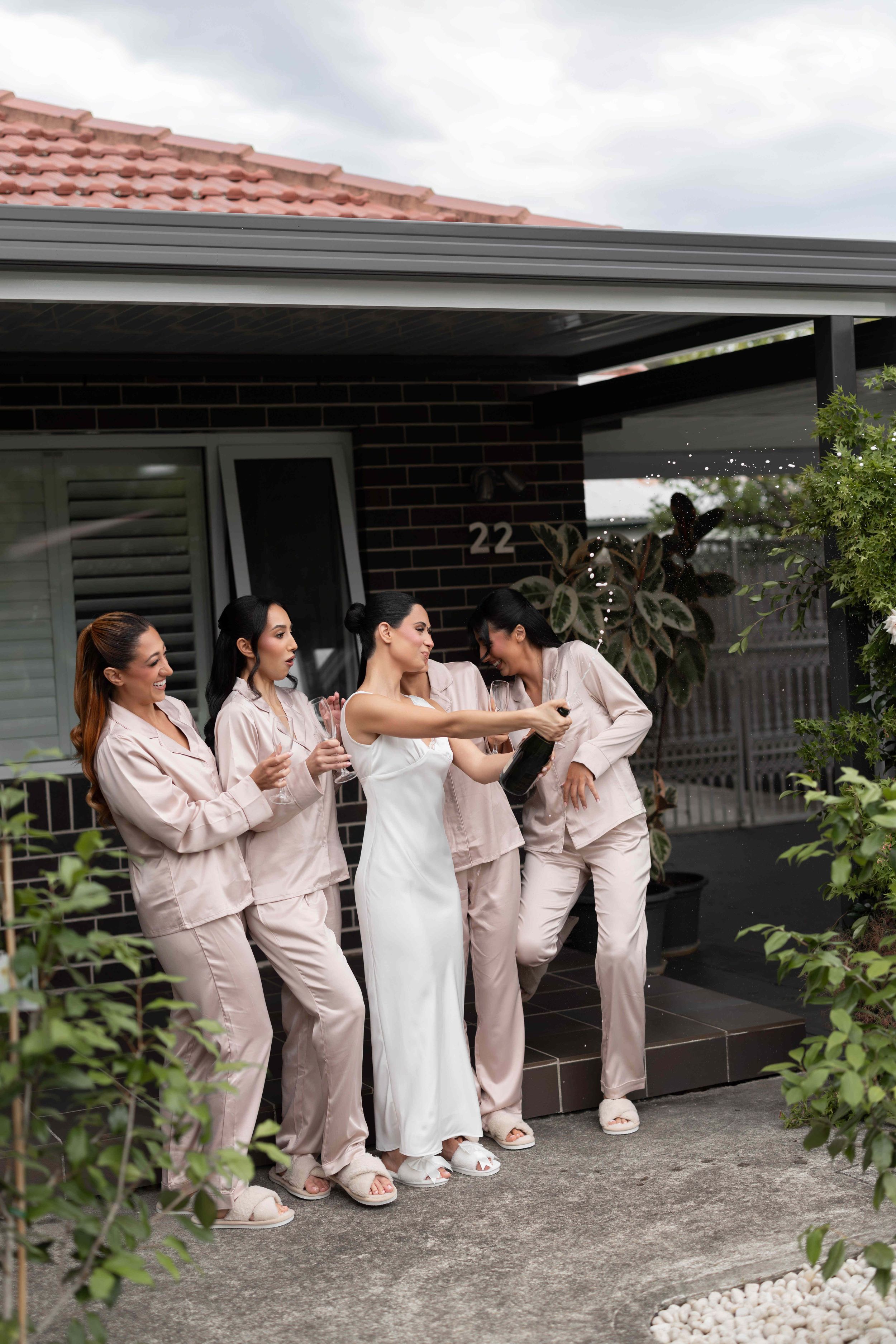 Bride in white dress with bridesmaids in pink pajamas celebrating with champagne outside a brick house.