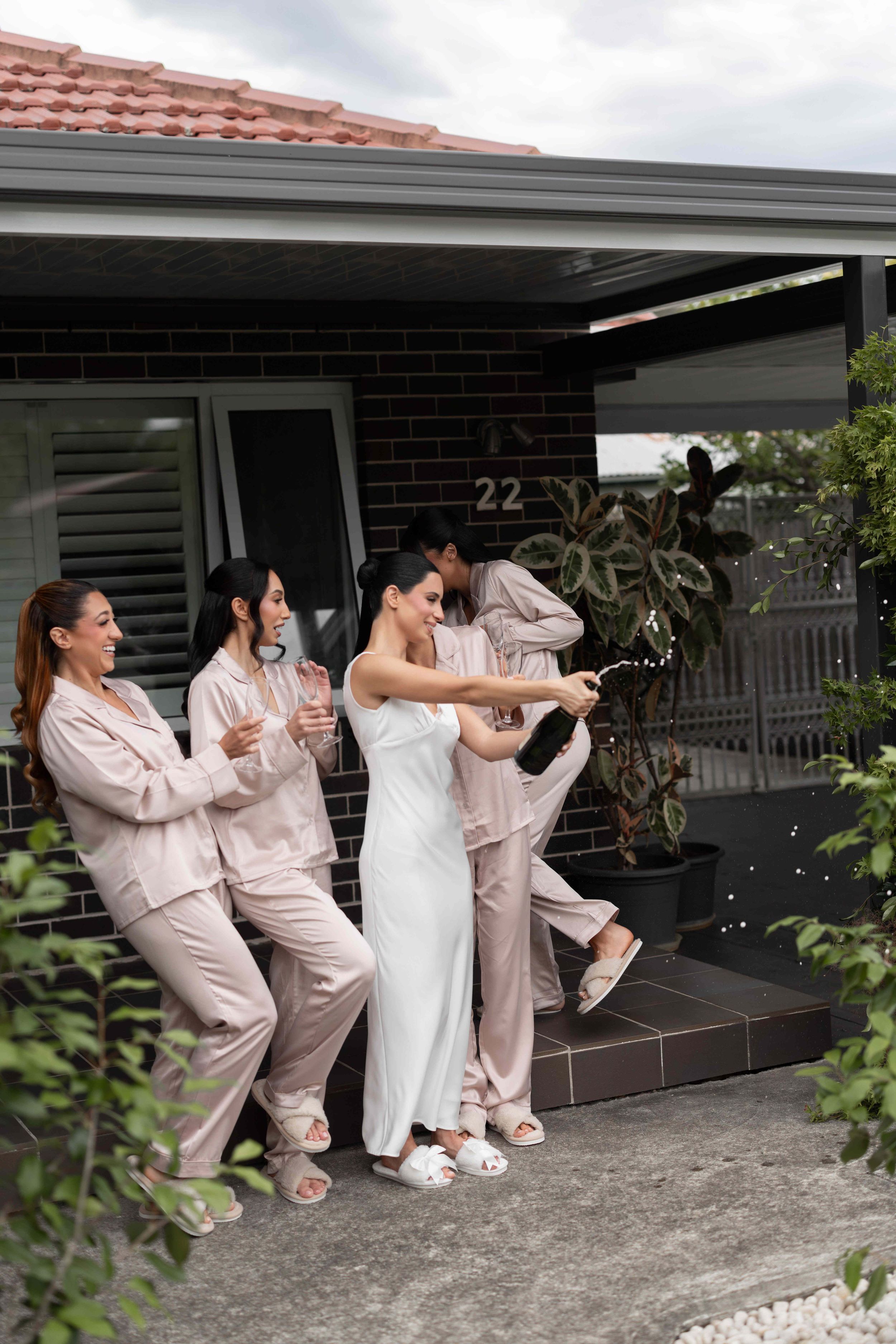 Bride in white dress pops champagne with bridesmaids in matching satin pajamas outside a brick home.