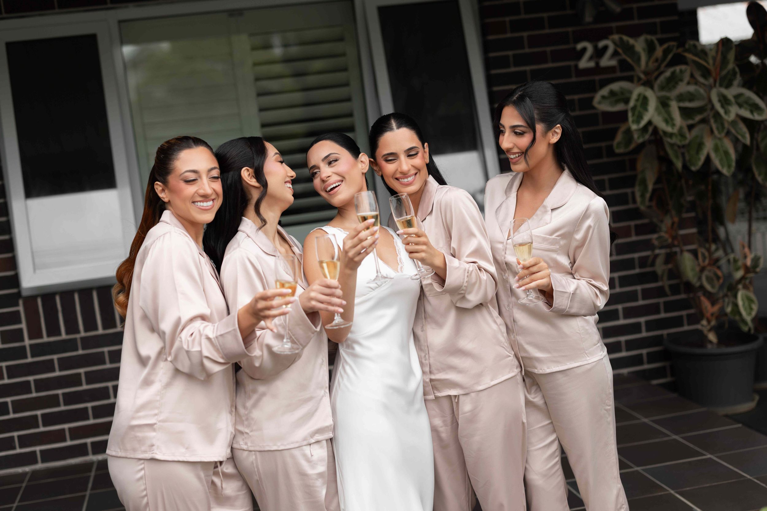 Bride in white gown with four bridesmaids in champagne satin pajamas toasting with champagne glasses outdoors.