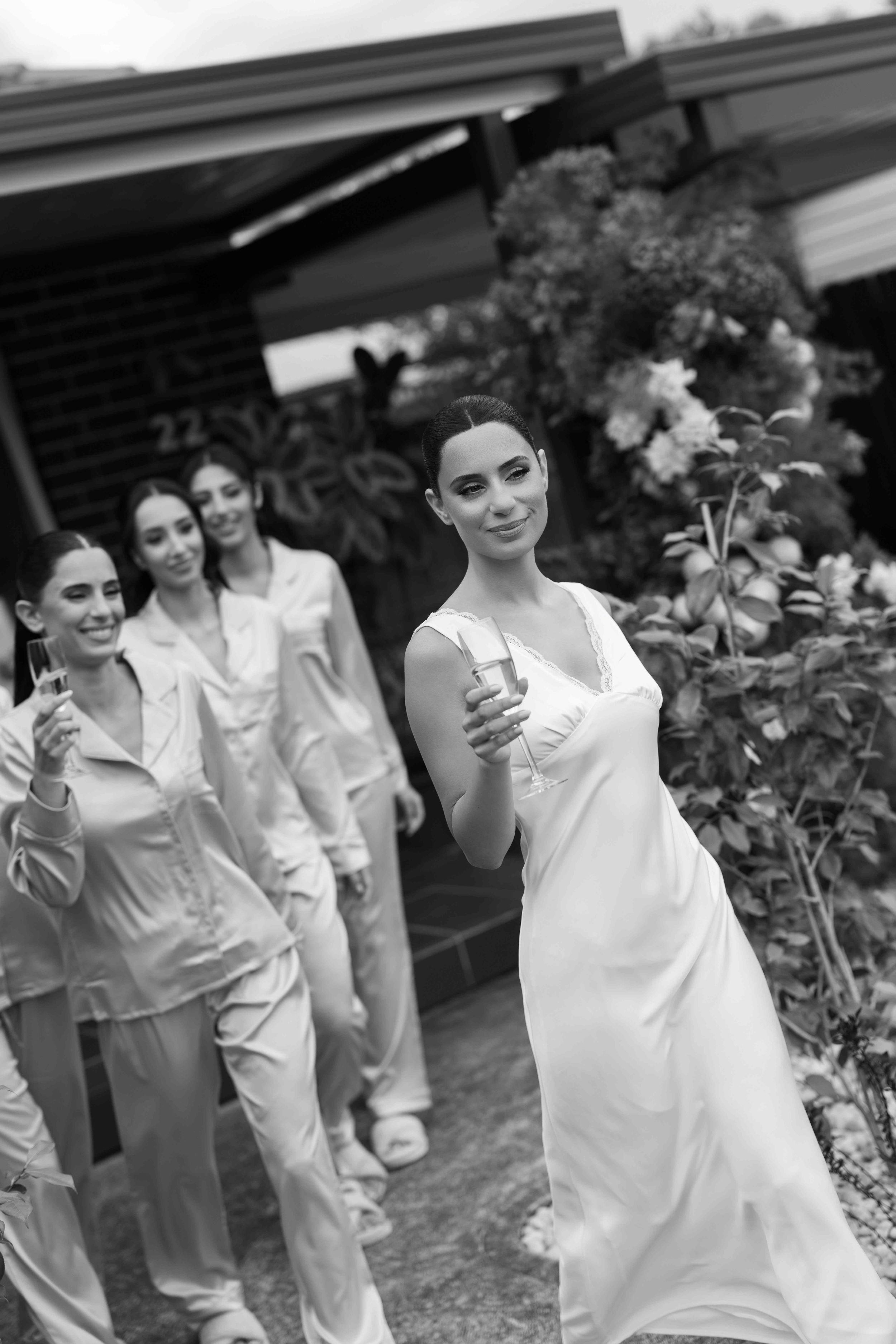 Black and white photo of a bride in a white slip dress holding champagne, walking with bridesmaids in satin pajamas outdoors.