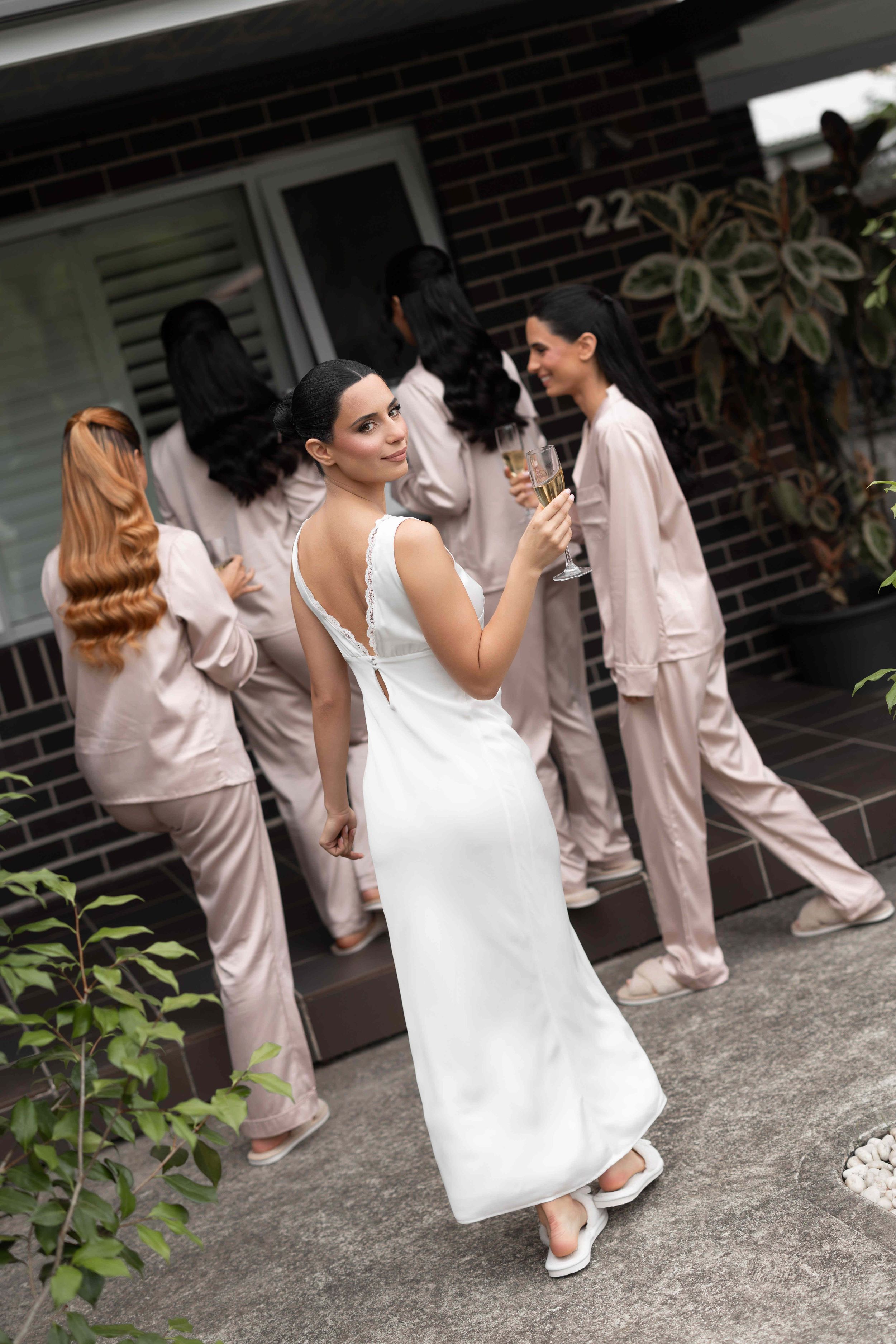 Bride in white gown looks back while bridesmaids in pink pajamas gather near brick building entrance.