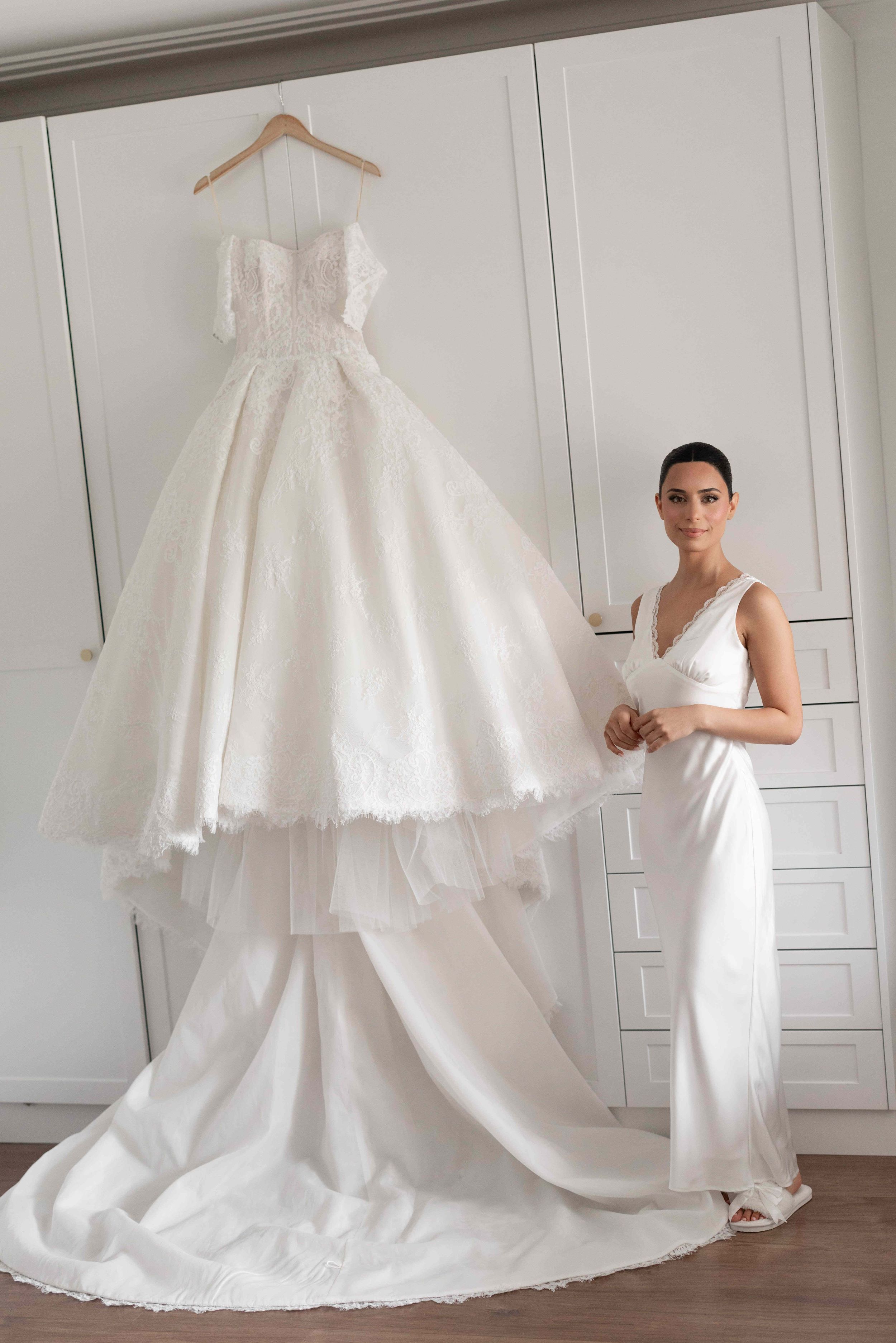 Bride in white satin gown admires elegant lace ballgown wedding dress on hanger in bright dressing room.