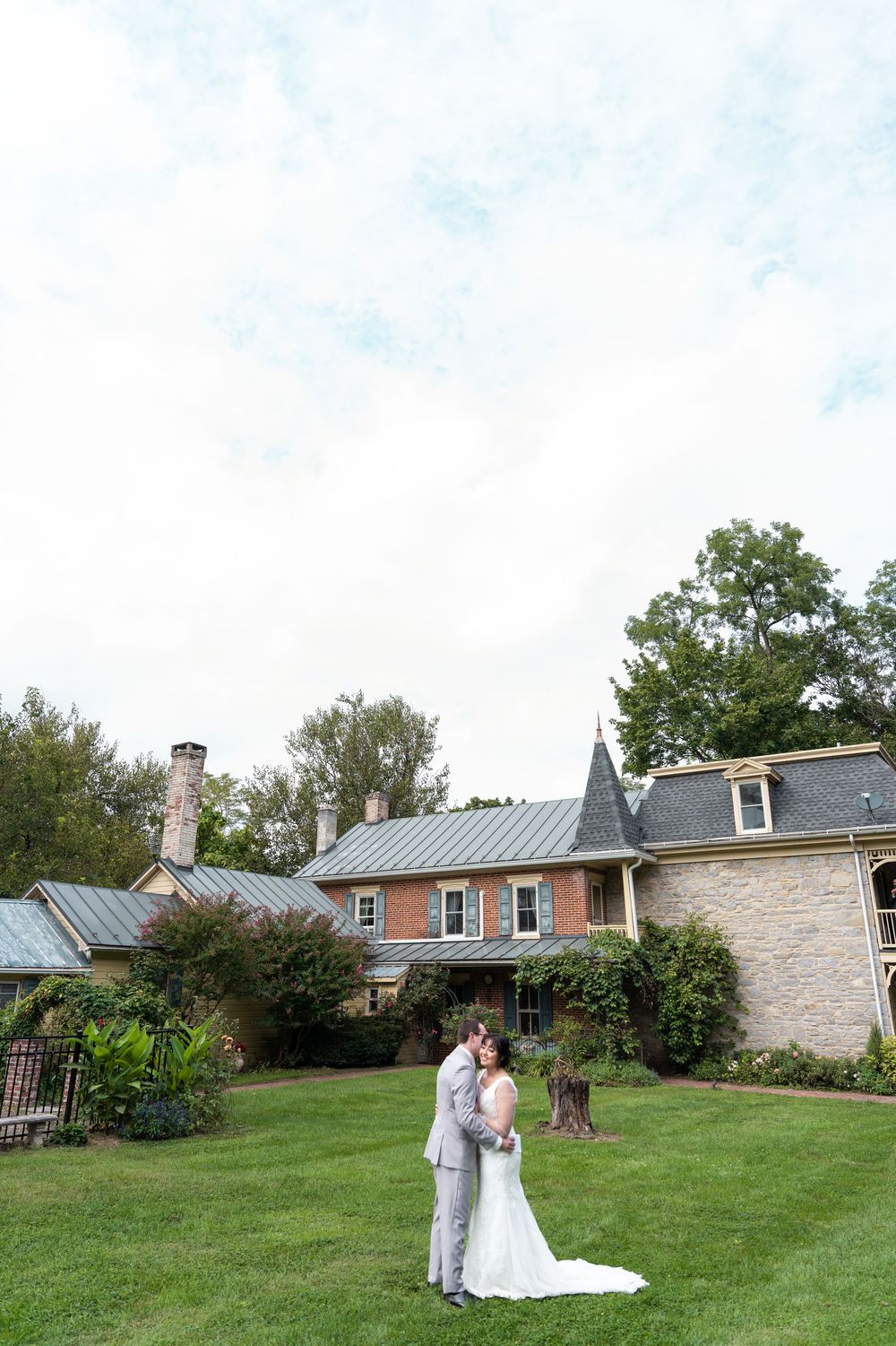 Bride and groom embrace on lush green lawn in front of elegant brick manor house on their wedding day.