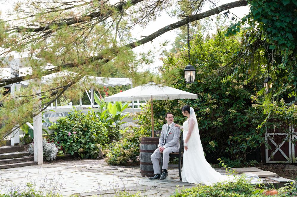 Bride and groom pose in a lush garden patio with white pergola, barrel seating, and lantern post on their wedding day.