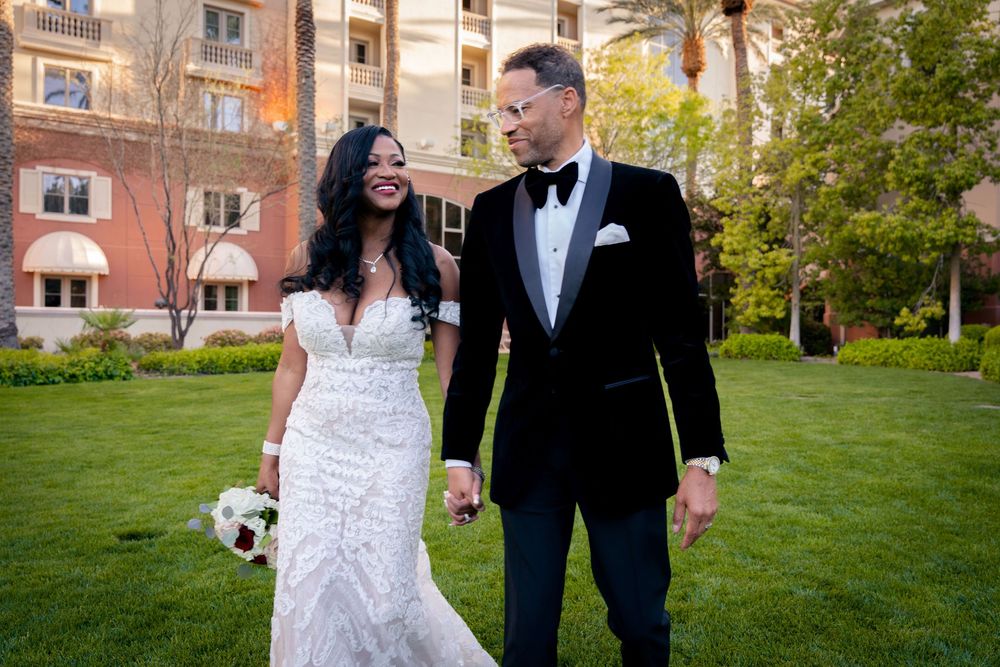Bride and groom walk hand-in-hand on green lawn, she in lace gown, he in black tuxedo, hotel backdrop.