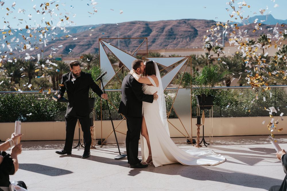 Couple shares first kiss at outdoor rooftop wedding ceremony with confetti and mountain views in the background.