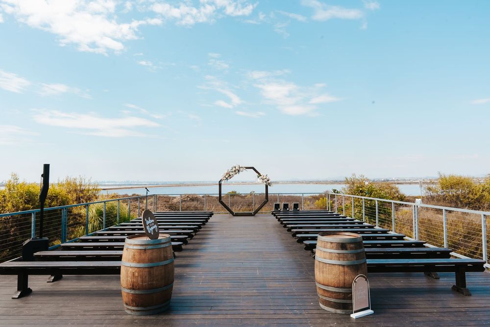 Outdoor wedding ceremony setup on a wooden deck overlooking the ocean with barrel decor and a floral arch.