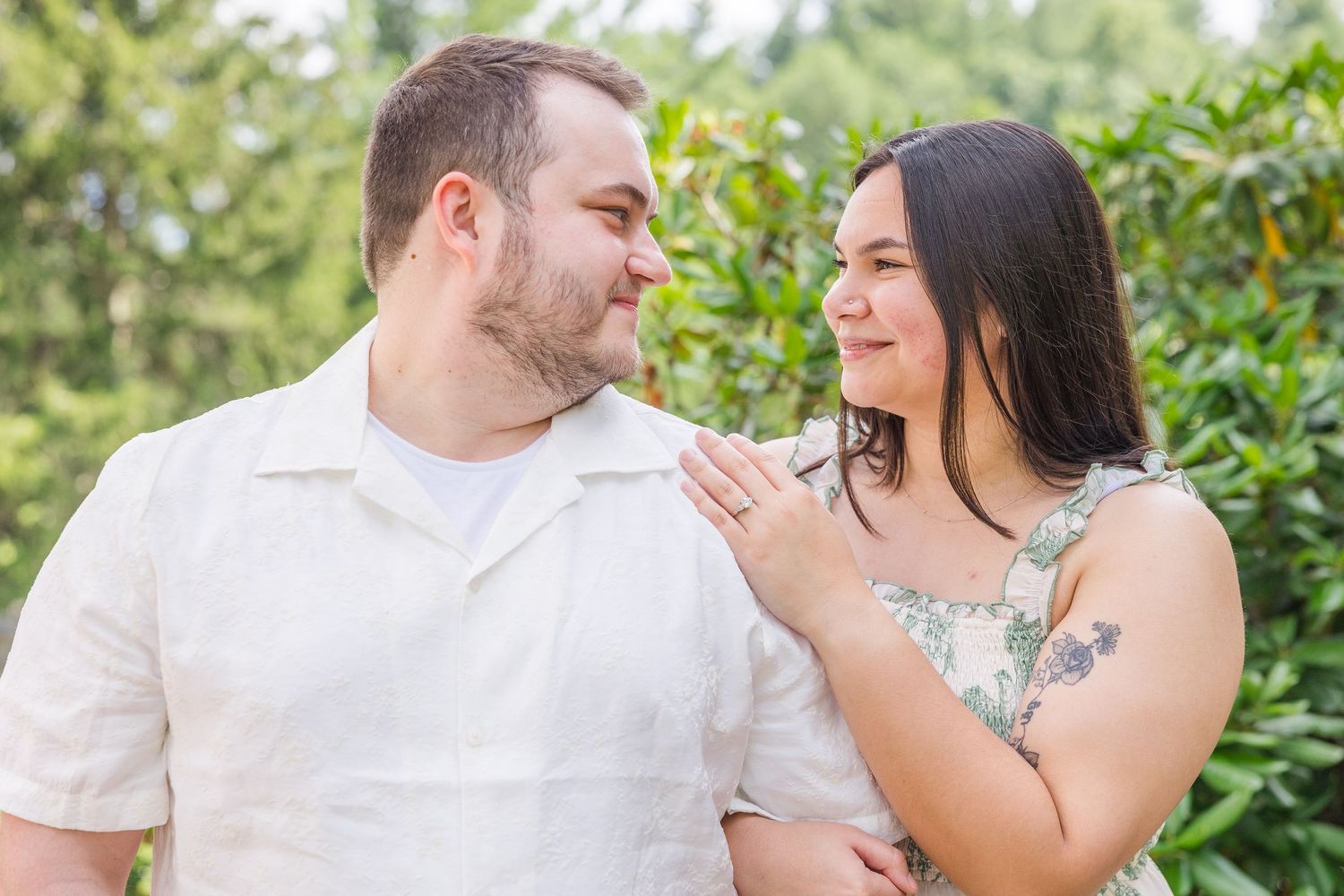 Couple sharing a tender moment outdoors, woman touching man's chin, smiling at each other with green foliage behind.