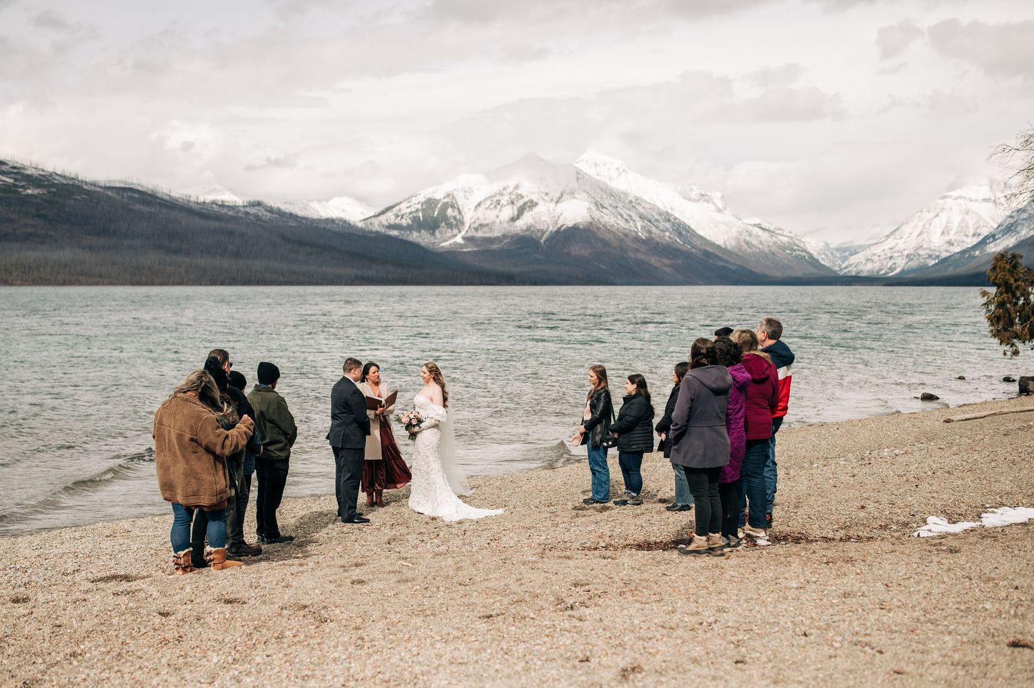 Bride and groom kissing on rocky lakeshore with stunning mountain backdrop under clear blue sky.