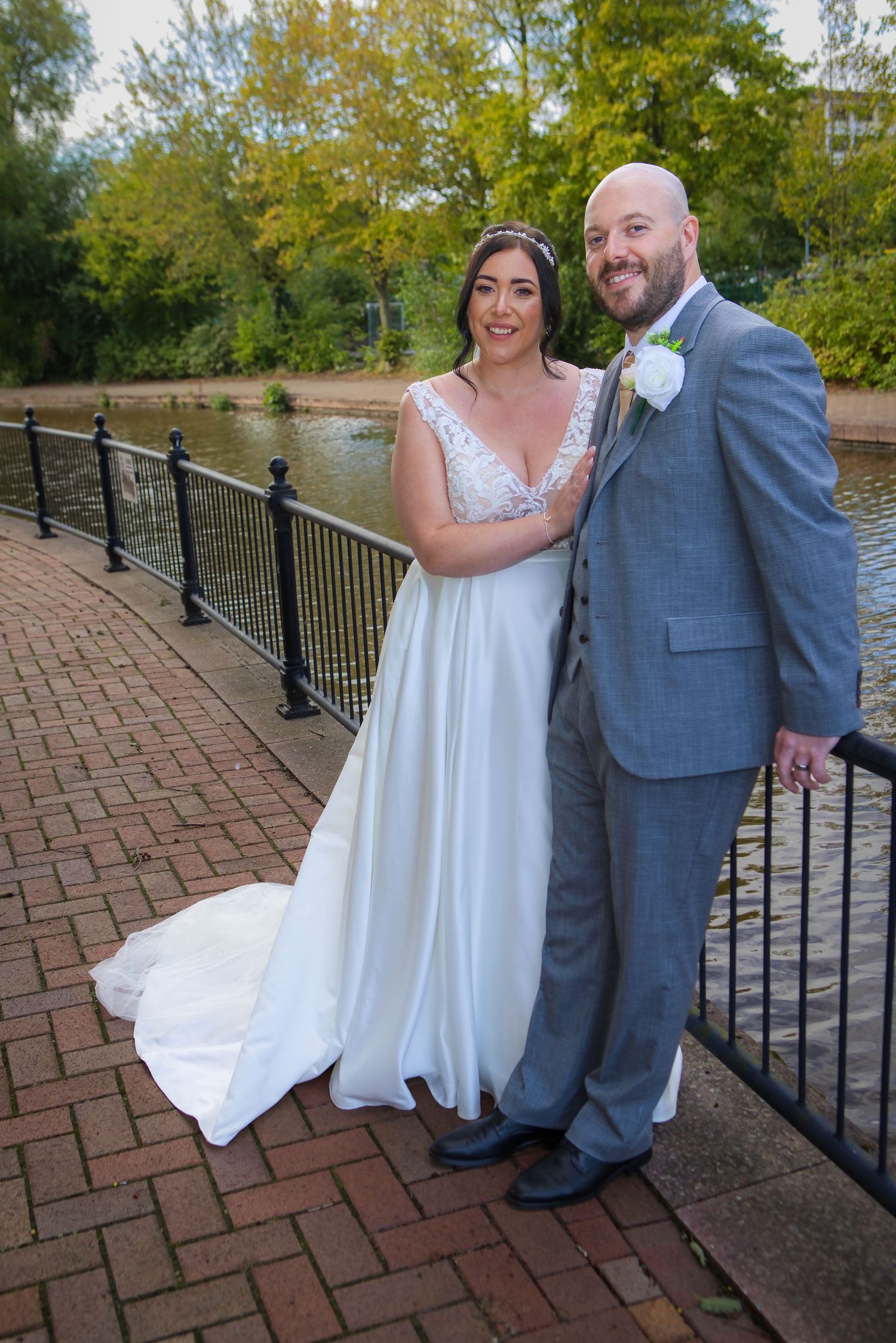 Bride in white gown and groom in gray suit pose together on a brick path by a railing near a pond.