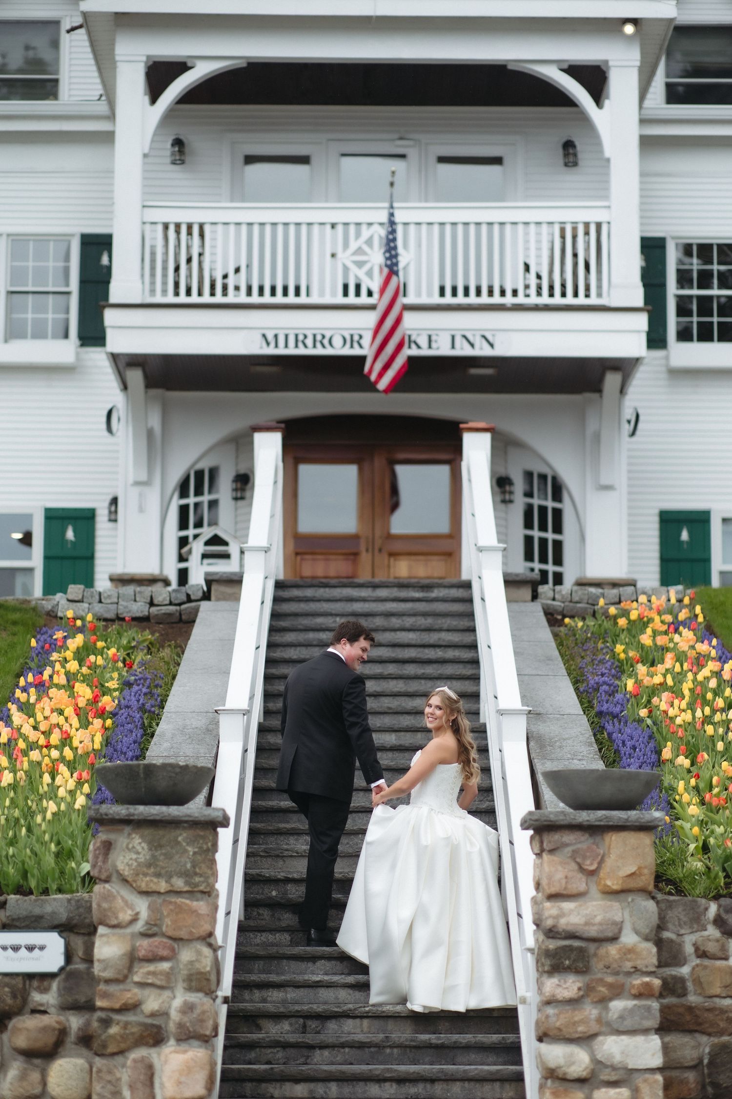 Bride and groom hold hands on stone steps at Mirror Lake Inn, surrounded by colorful tulips and an American flag.