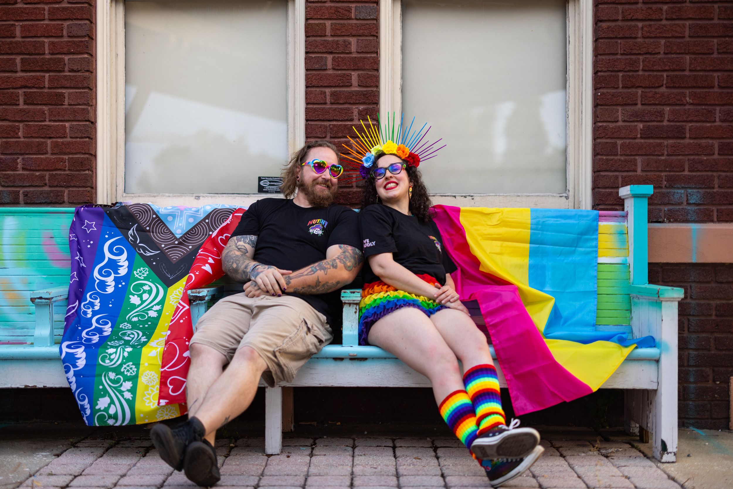 Two people sitting on a bench draped in rainbow Pride flags, smiling, wearing colorful Pride accessories outdoors.