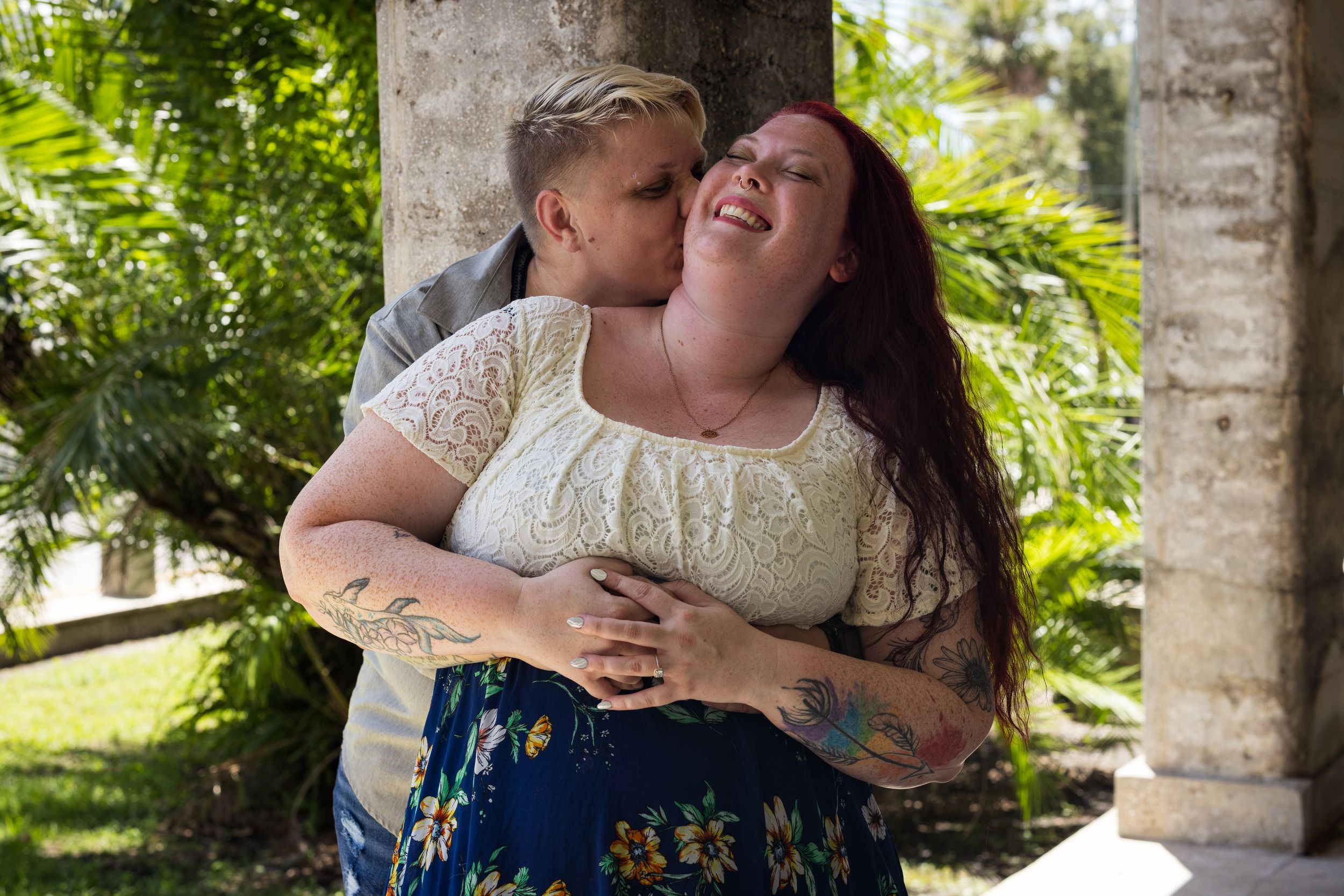 Couple embracing outdoors, woman laughing joyfully as man kisses her cheek near tropical palm columns.