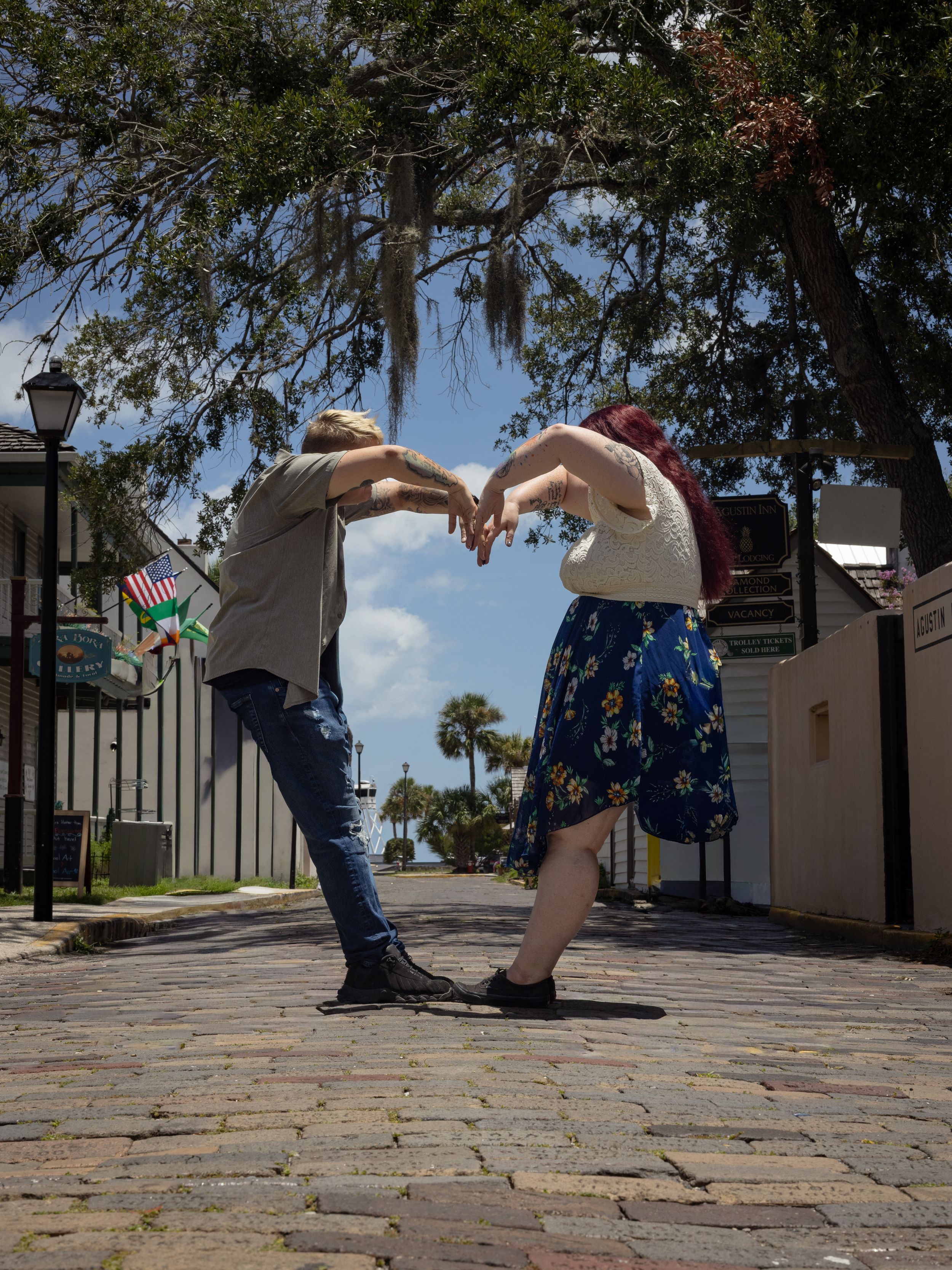 Couple forming a heart shape with their hands on a cobblestone street lined with trees and flags.