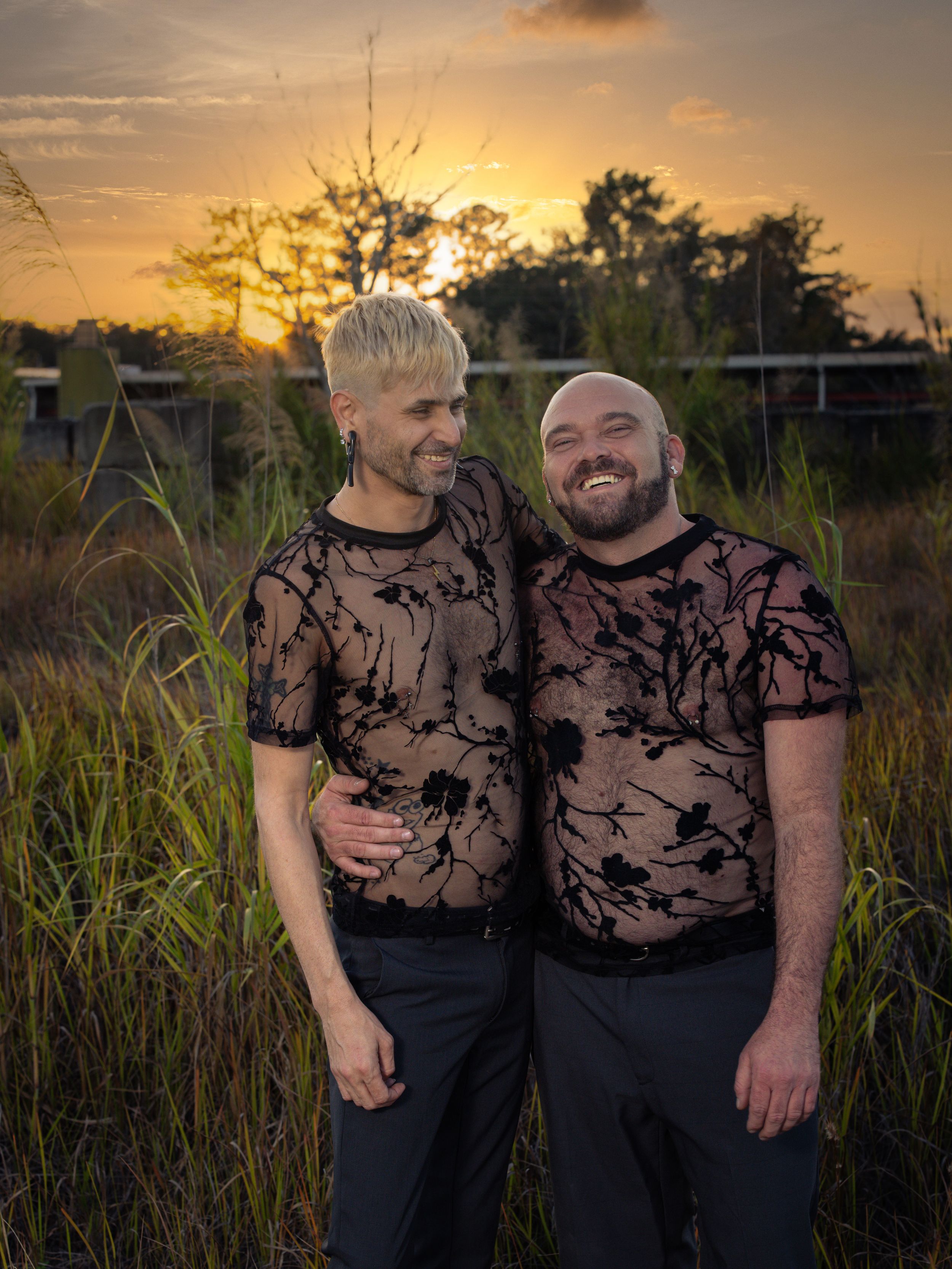 Two people in matching black floral sheer tops embrace smiling at sunset in a field of tall grass.