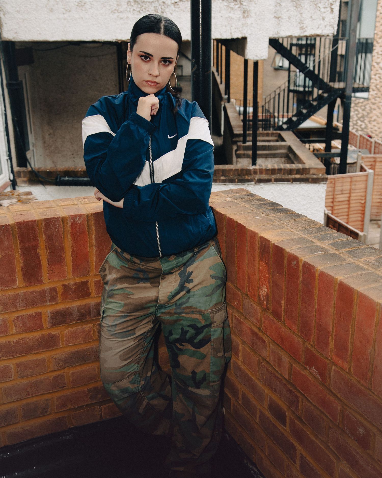 Woman in Nike jacket and camo cargo pants posing against brick wall outdoors.