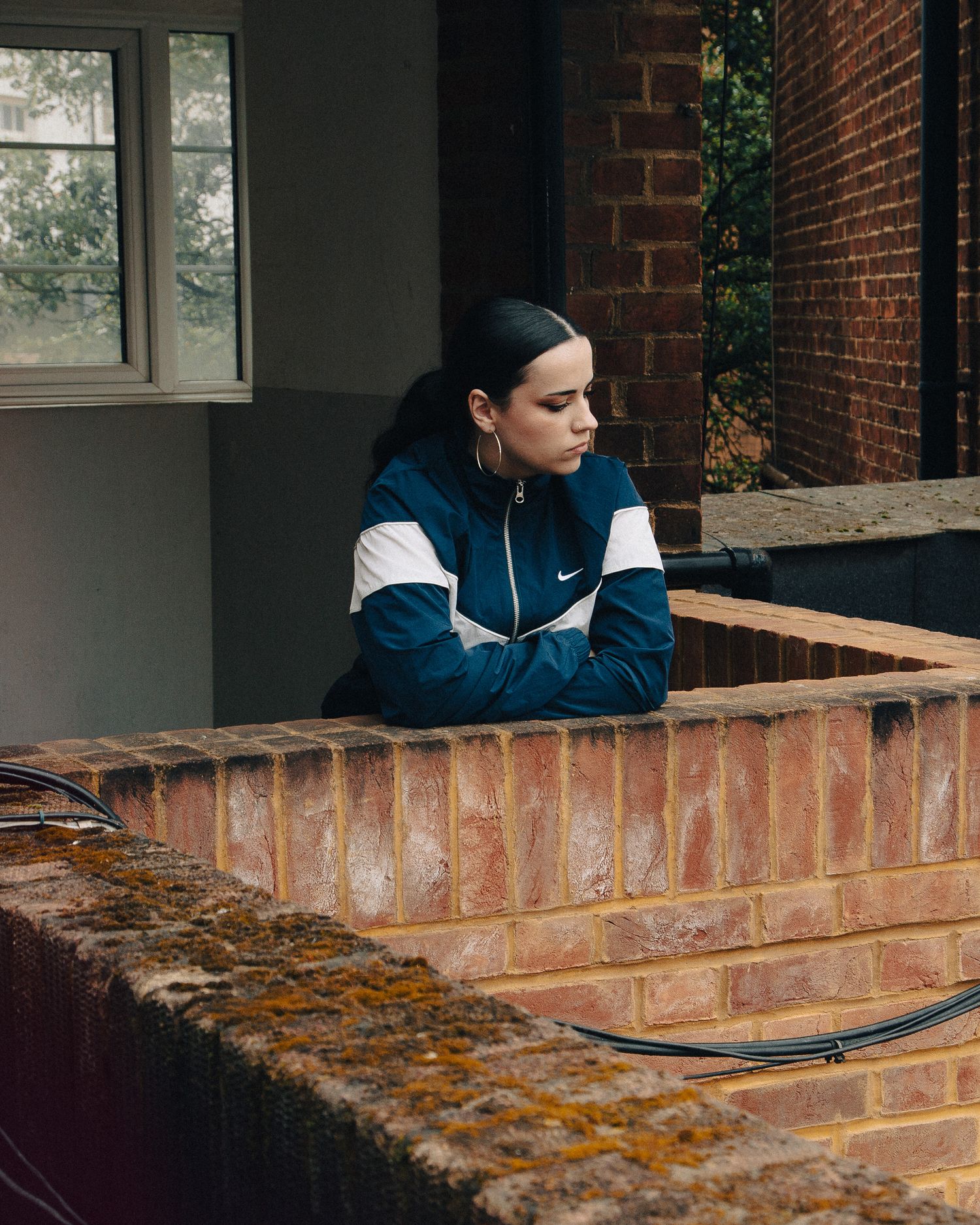 Young woman in a teal Nike track jacket leaning on a brick wall outdoors near a brick building.