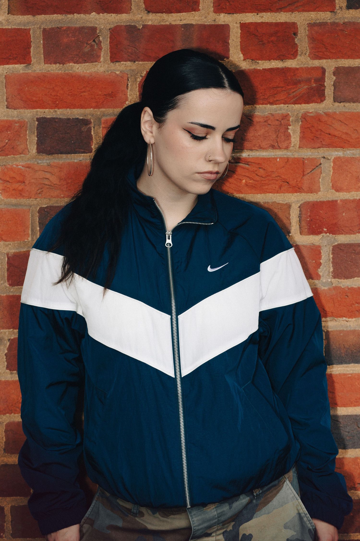 Young woman in navy blue and white Nike windbreaker jacket posing against a red brick wall.