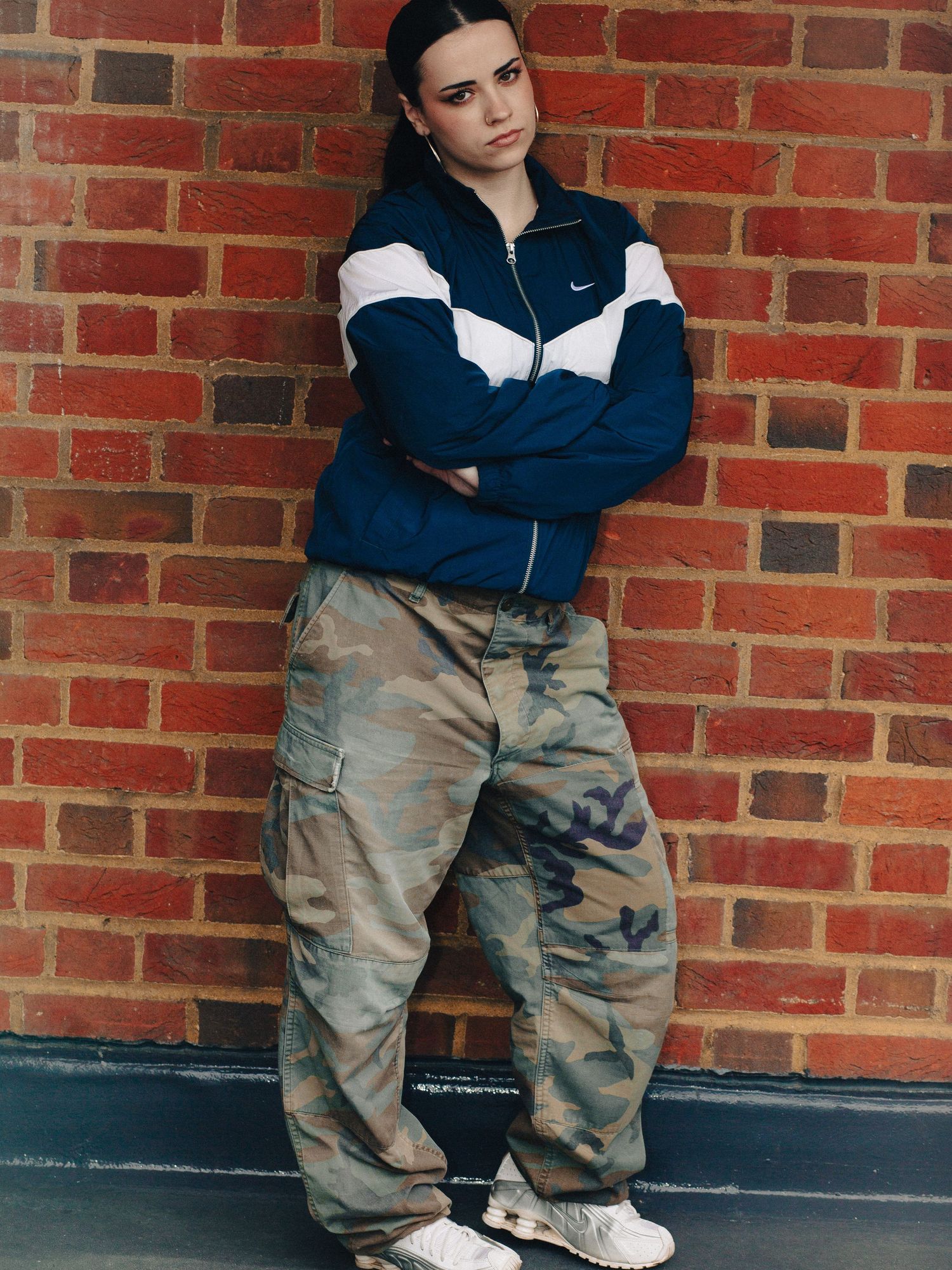Young woman in Nike track jacket and camo cargo pants leaning against brick wall with arms crossed.
