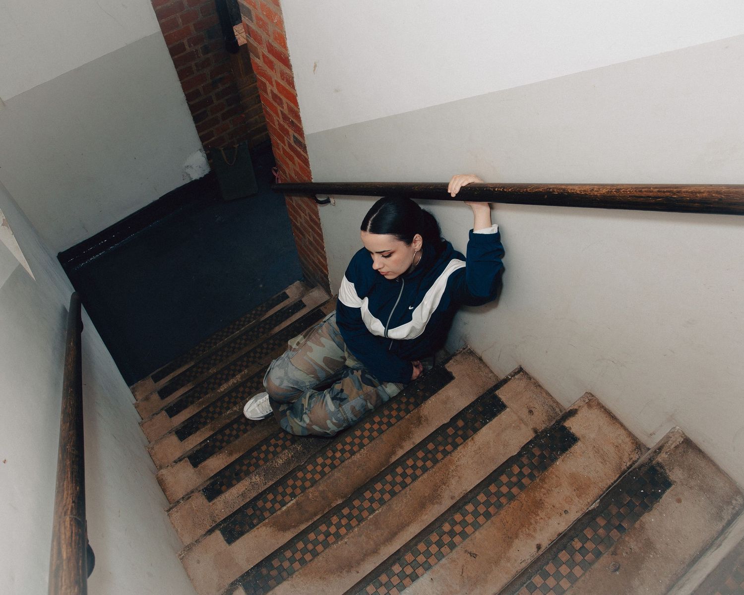 Young woman sitting on worn wooden stairs in an old building stairwell, holding a railing overhead.