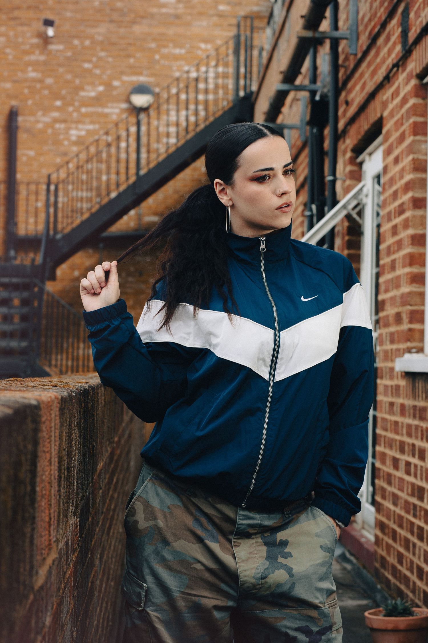 Young woman in navy Nike track jacket and camo pants poses outdoors near brick building with metal stairs.