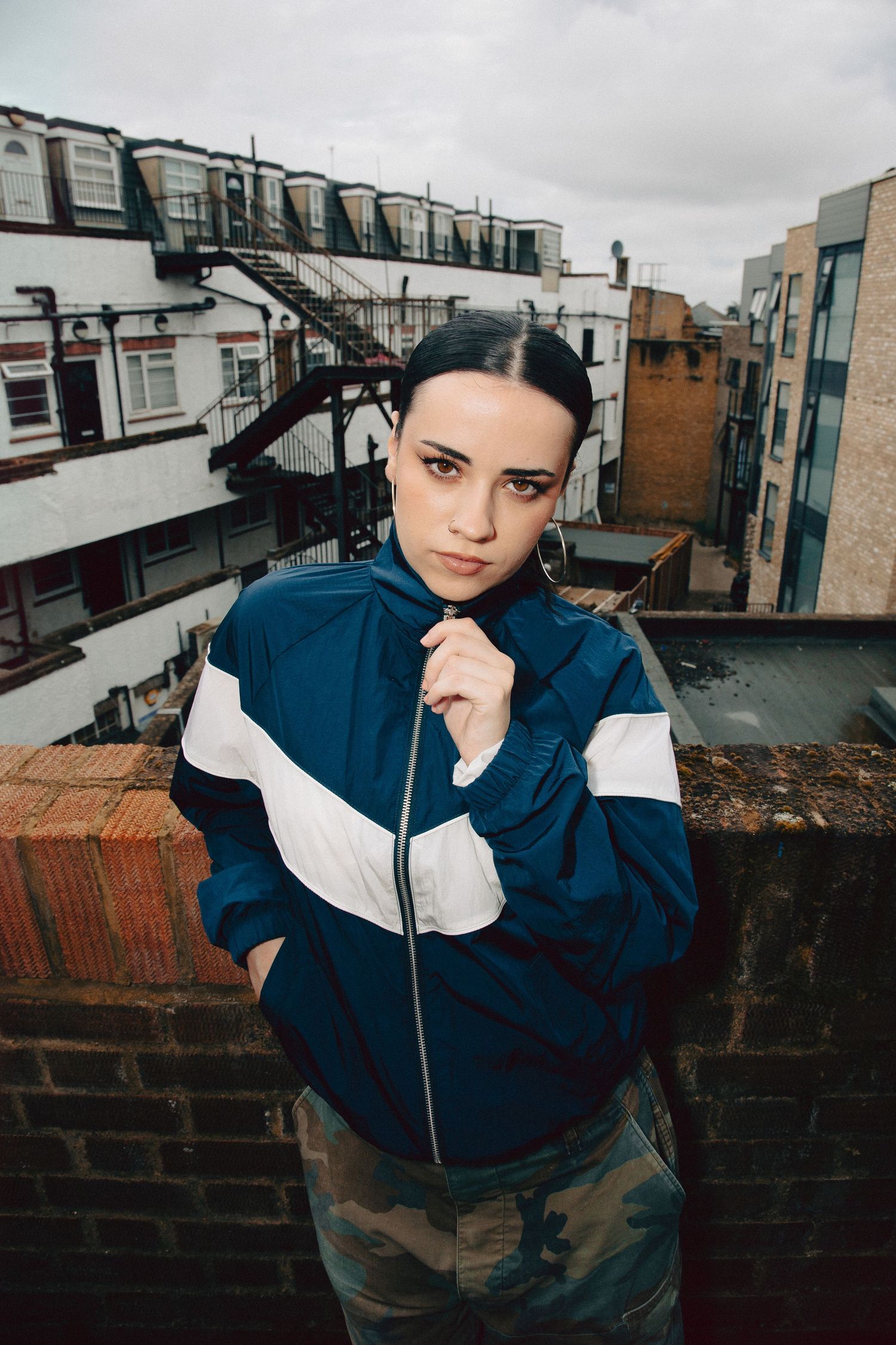 Young woman in blue tracksuit jacket poses on urban rooftop with city buildings in background.