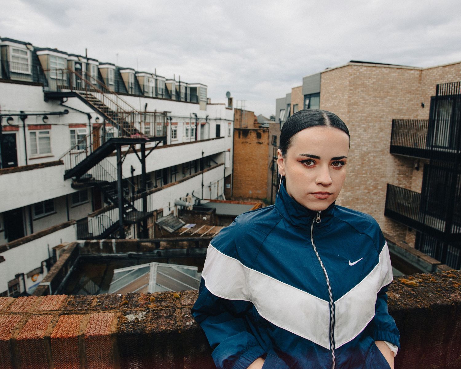 Young woman in Nike tracksuit jacket poses on urban rooftop with city buildings in background.