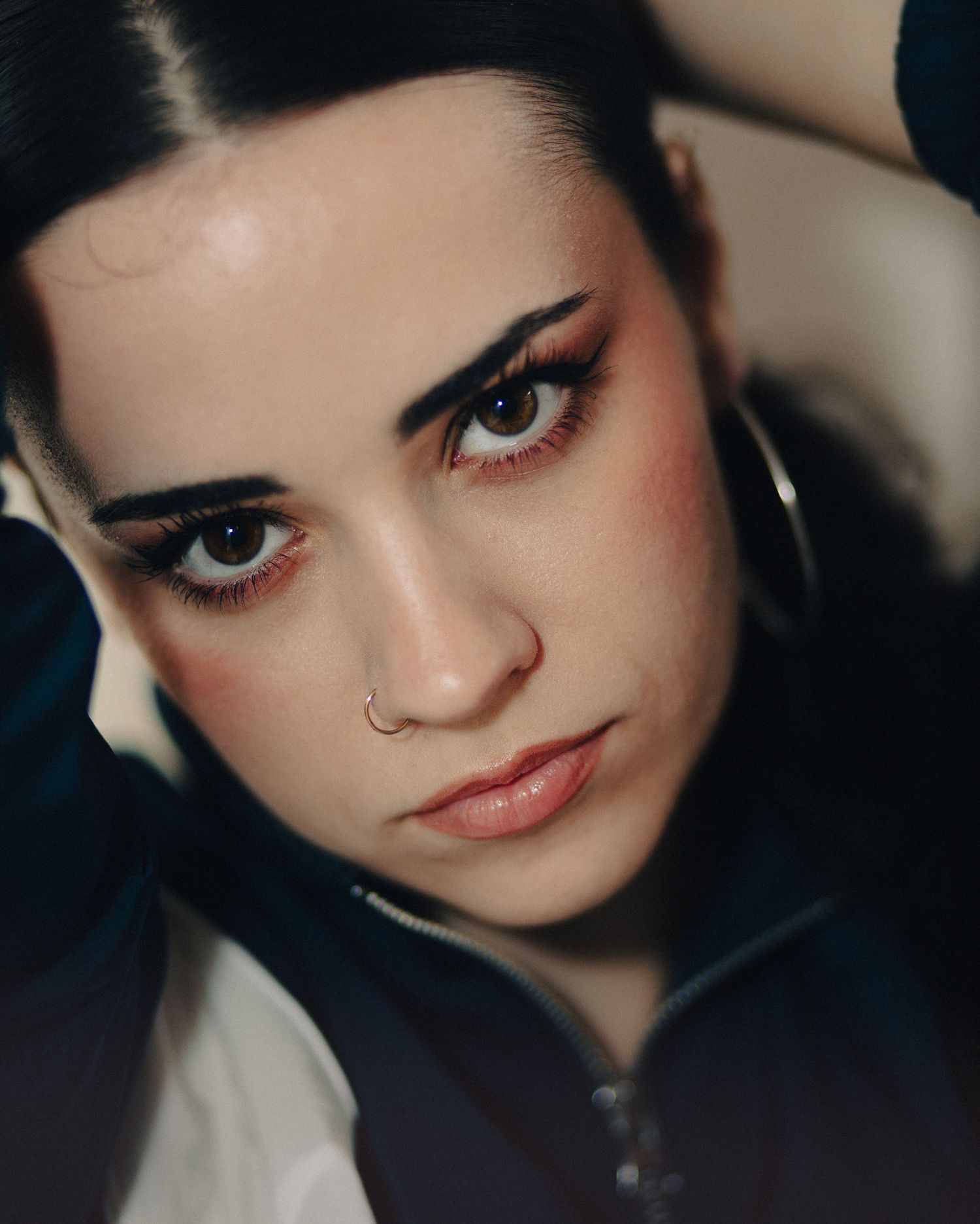 Close-up portrait of a young woman with bold eyeliner, hoop earrings, and a nose ring against a dark background.