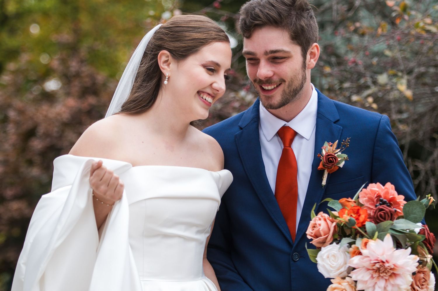 A smiling bride in a white dress gazes at her groom wearing a floral boutonniere in warm golden sunset light.