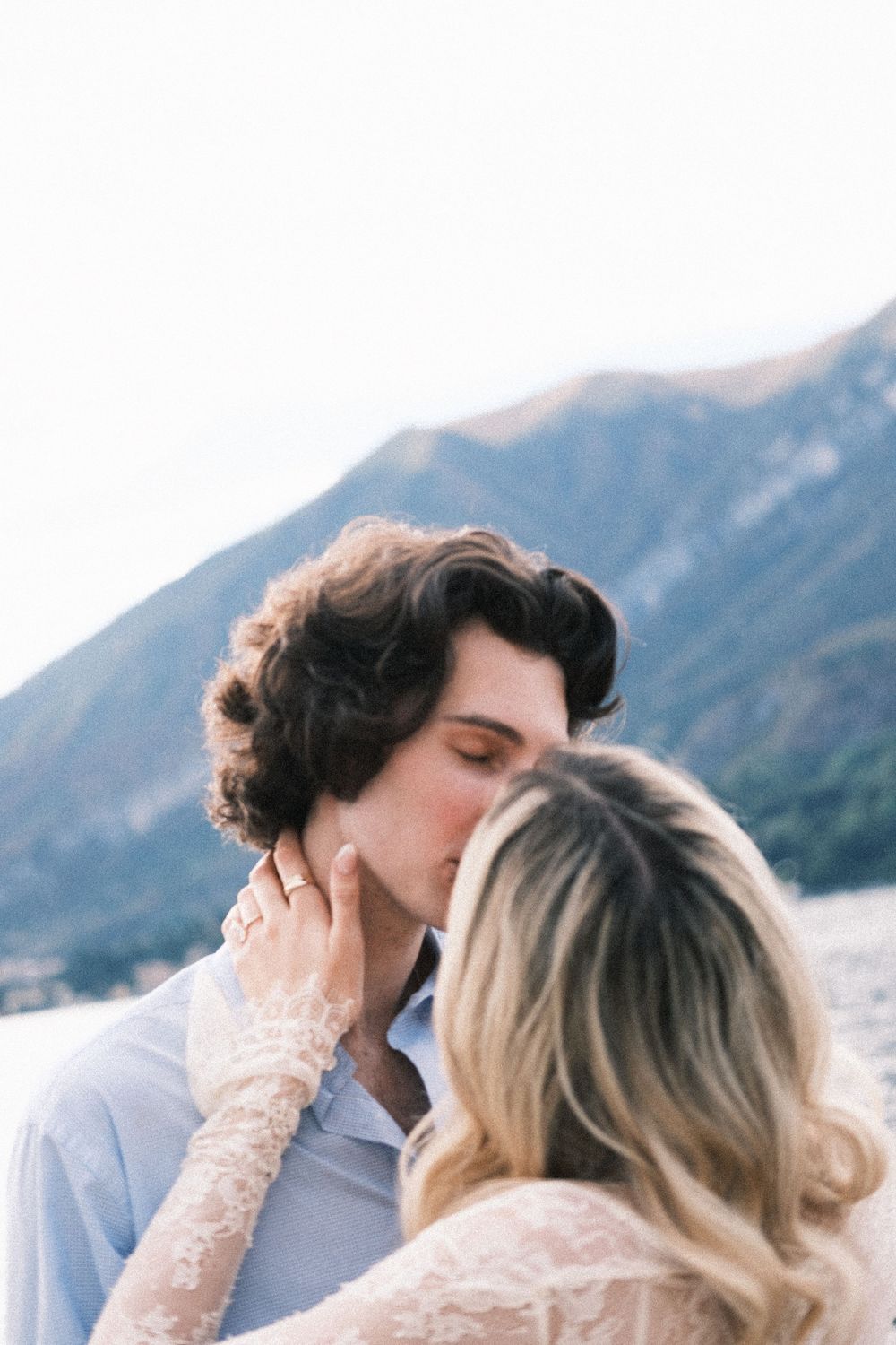 A couple shares a romantic kiss with scenic mountain lake backdrop, bride in lace dress tenderly holds groom's face.