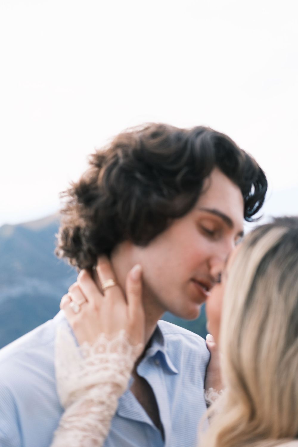 Romantic couple sharing a tender kiss outdoors, woman wearing lace sleeve and ring, mountains in background.