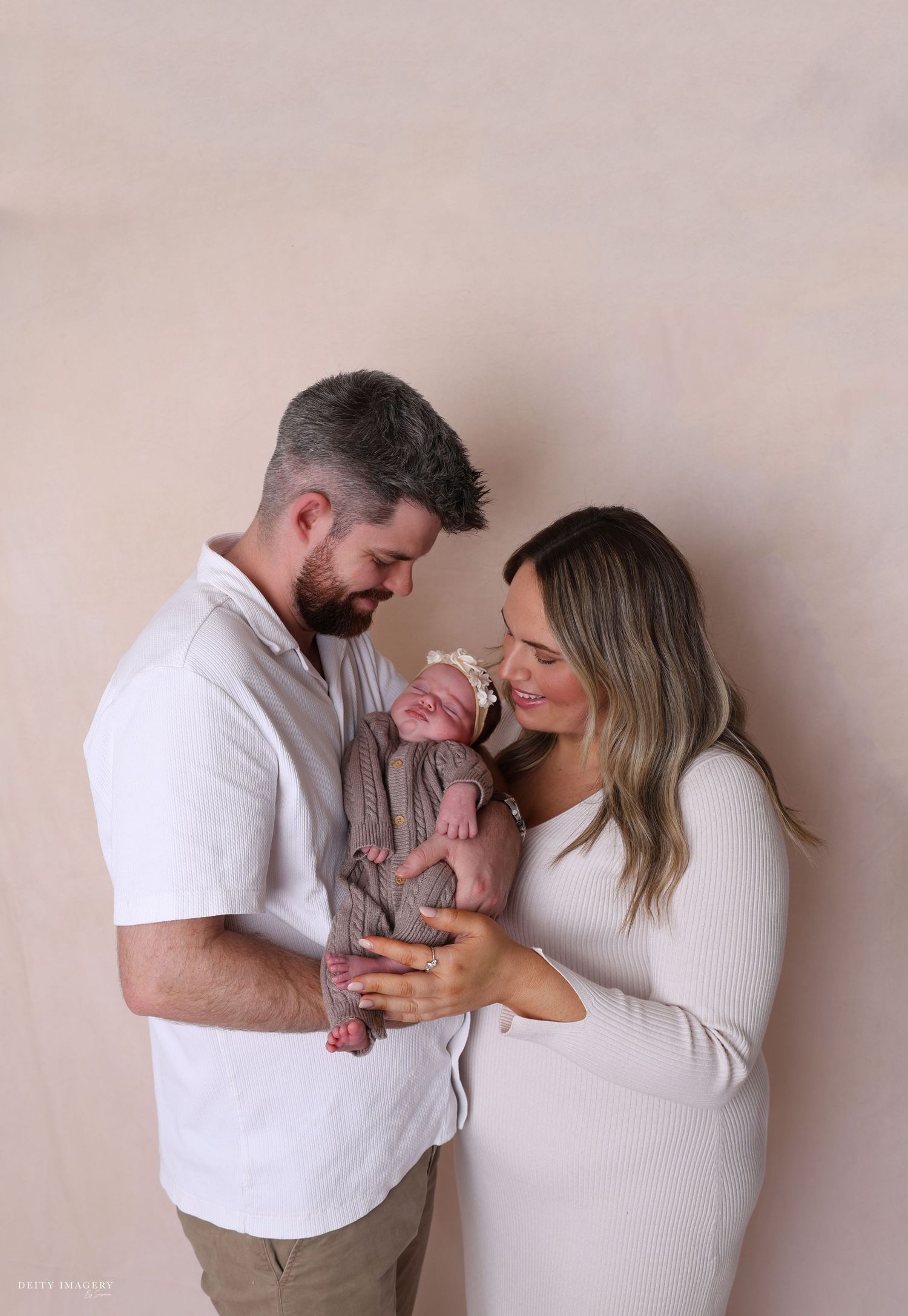 A couple in white outfits lovingly holds their newborn baby against a soft beige backdrop.