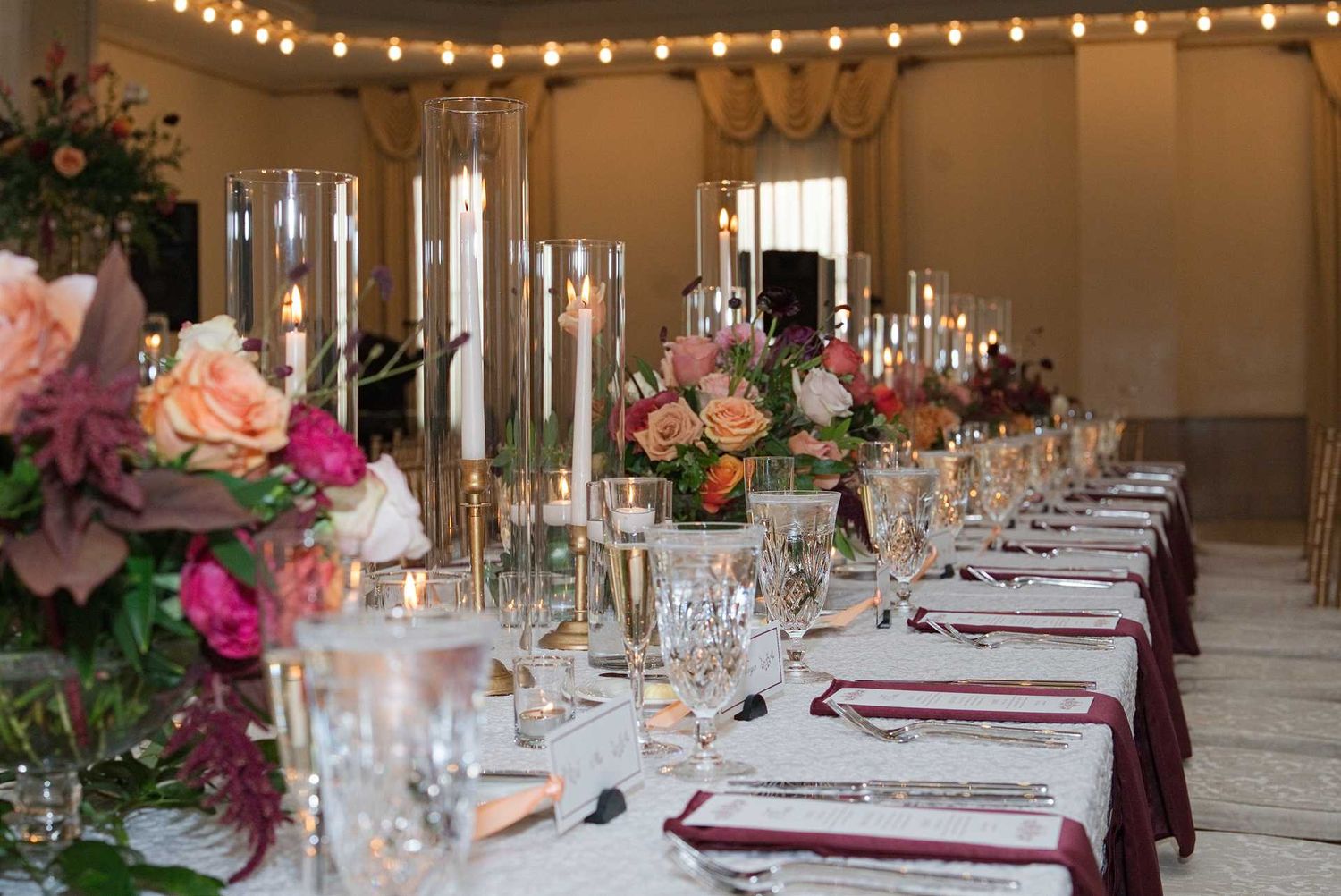 Elegant wedding reception table with tall glass candle holders, lush floral centerpieces, crystal glassware, and burgundy napkins