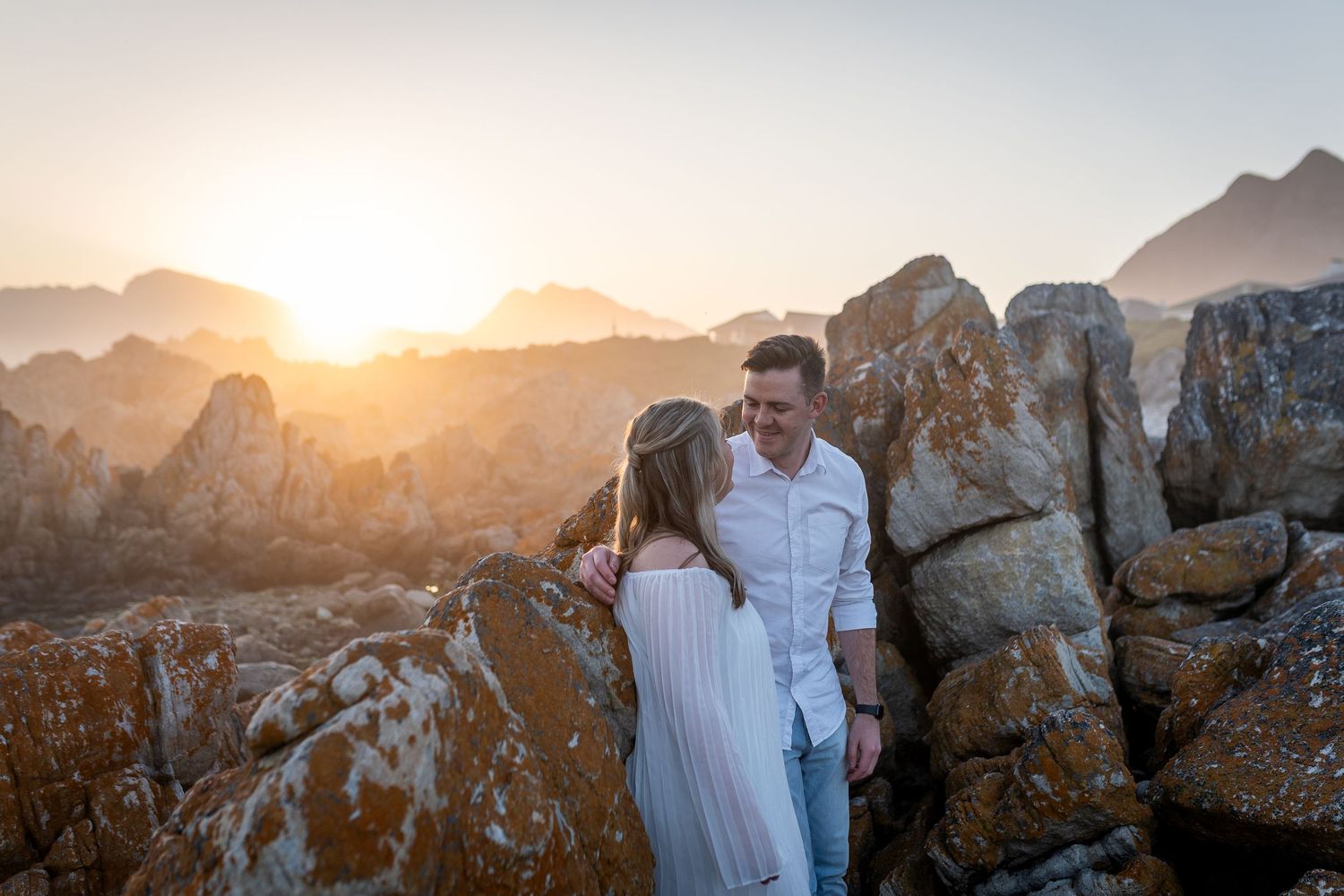 Couple embracing among rocky boulders at golden sunset, romantic outdoor engagement photo session.