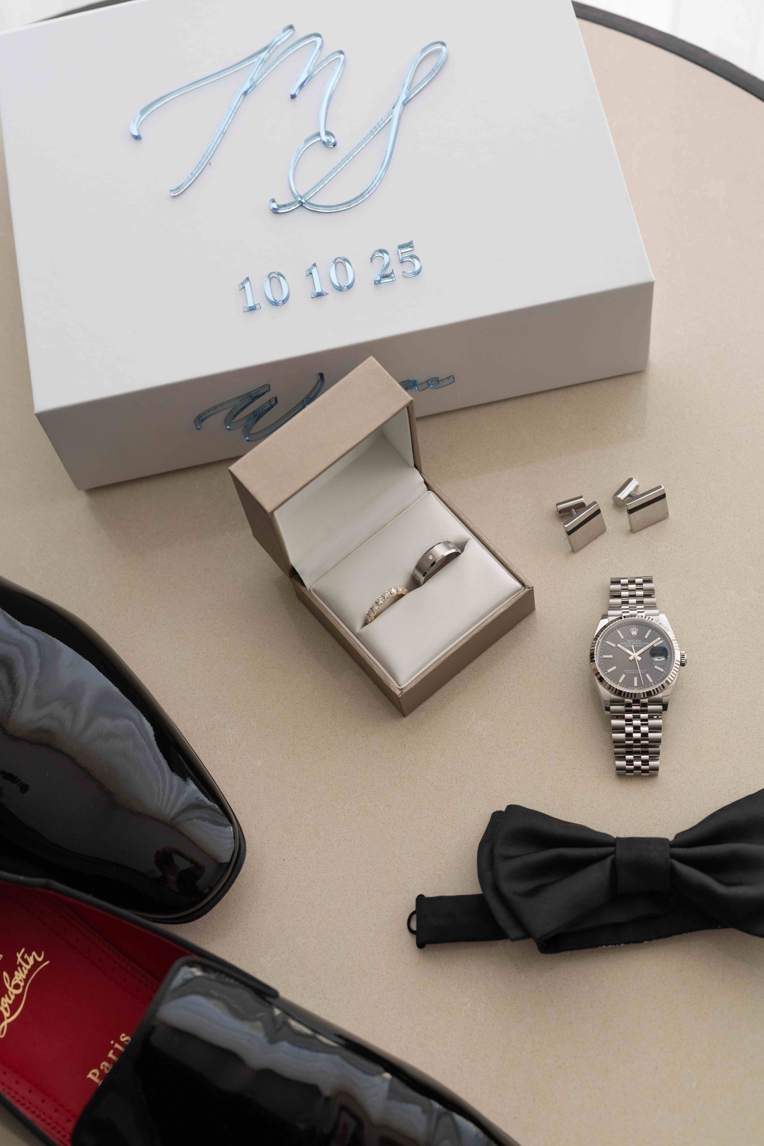 Elegant groom flat lay with Rolex watch, ring box, cufflinks, bow tie, Louboutin shoes, and monogrammed gift box dated 10.10.25.