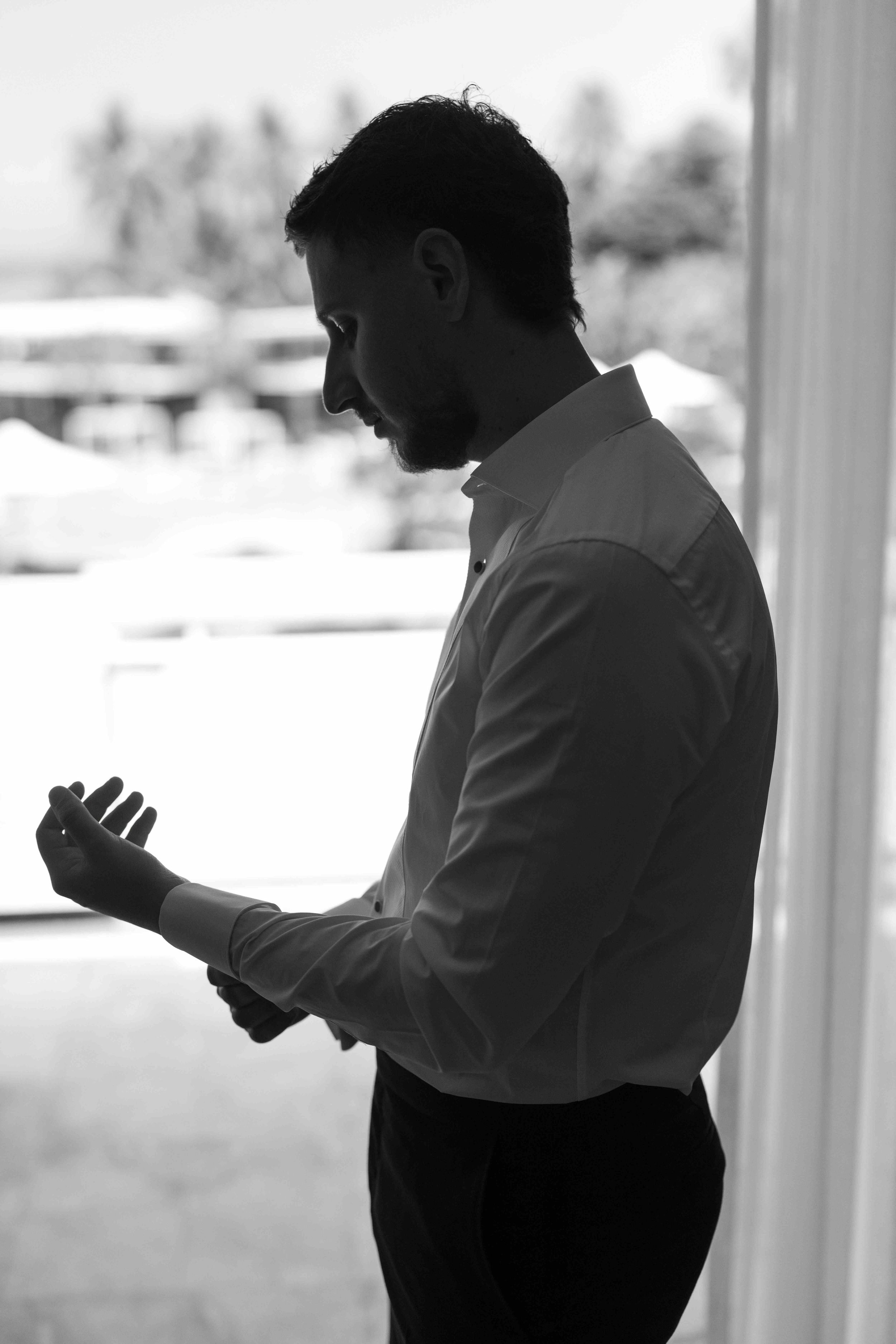 Black and white photo of a man in a dress shirt adjusting his cufflinks by a bright window.