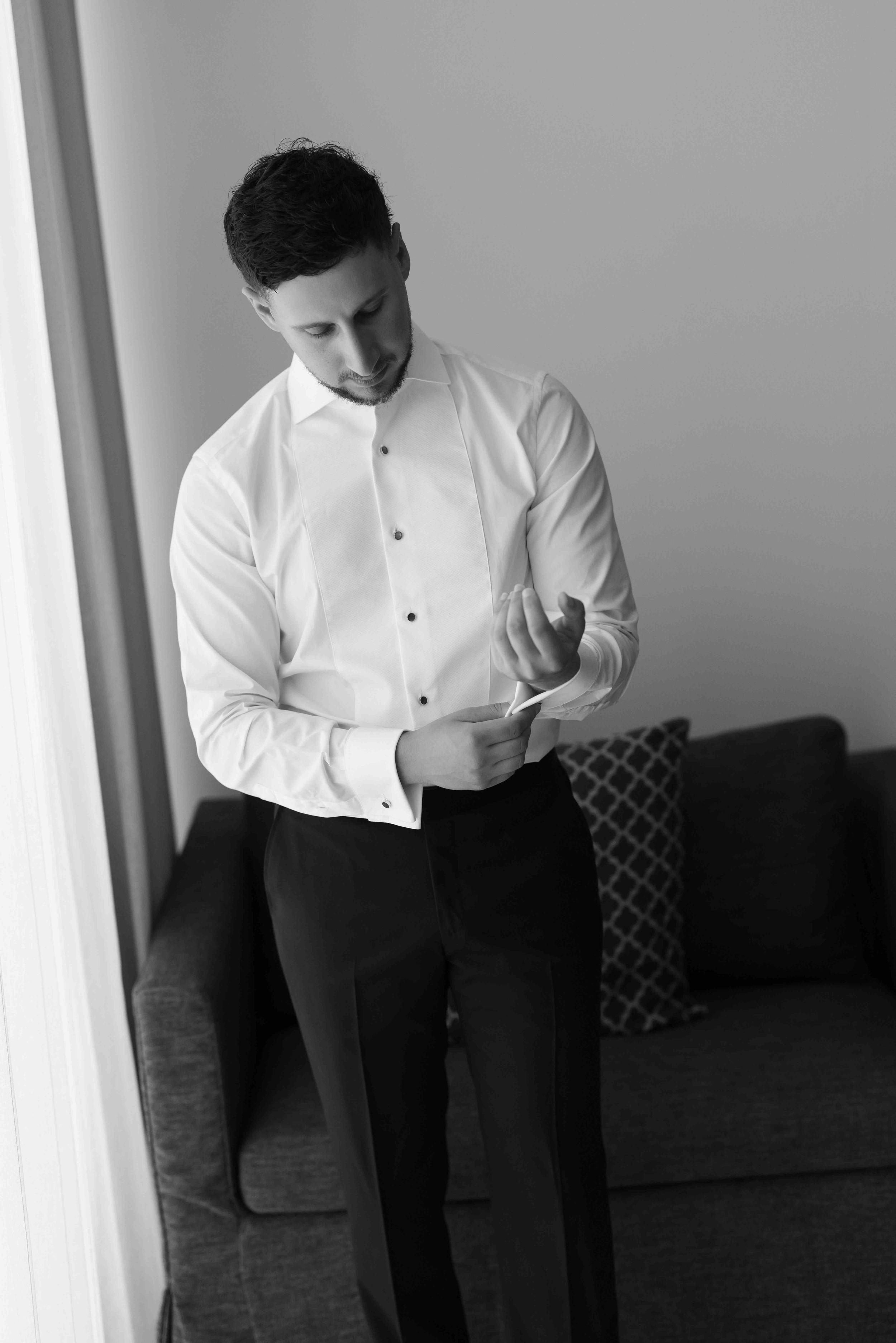 Black and white photo of a groom in a white dress shirt adjusting his cufflinks before his wedding ceremony.