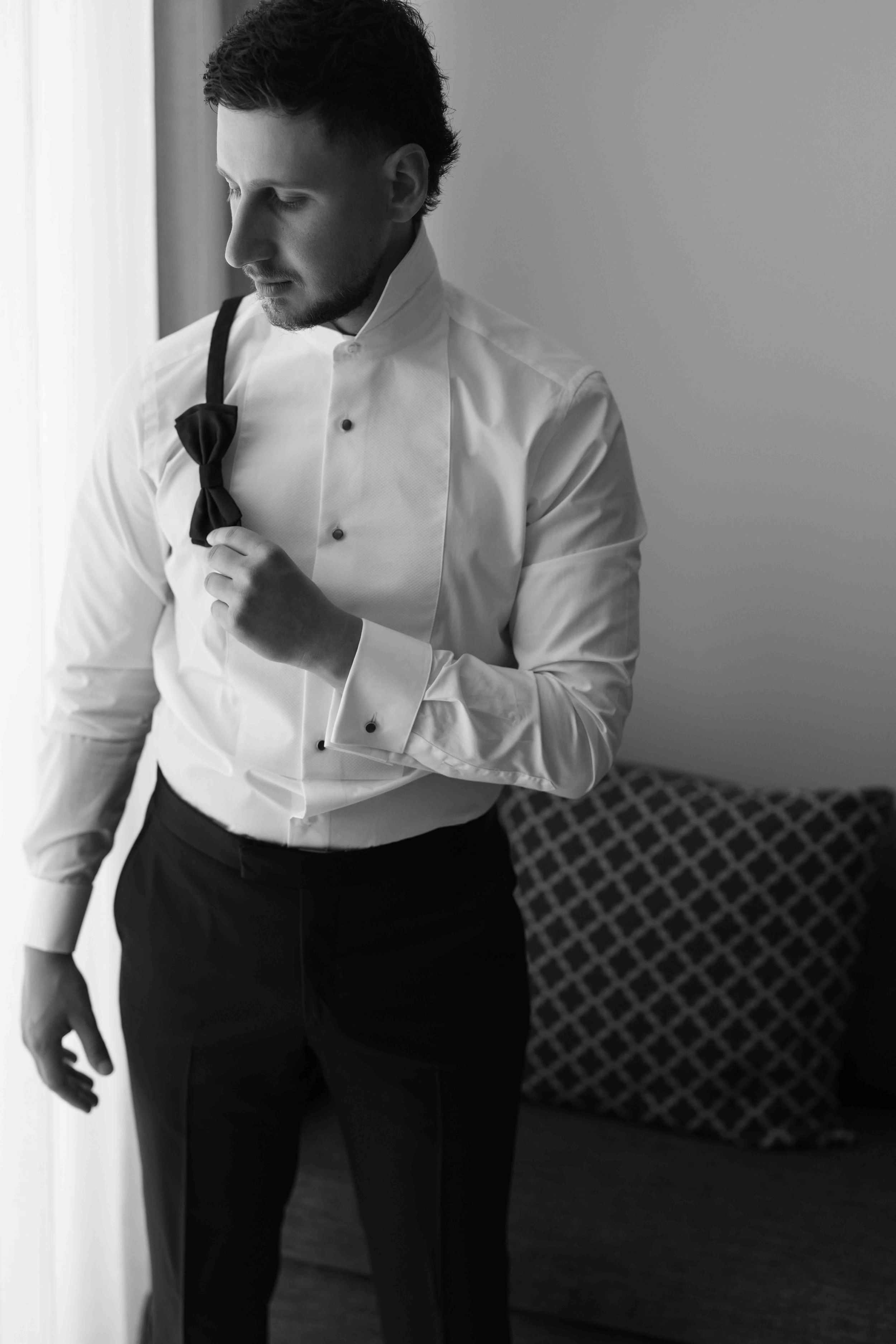 Black and white photo of a groom in a white tuxedo shirt adjusting his bow tie before the wedding ceremony.