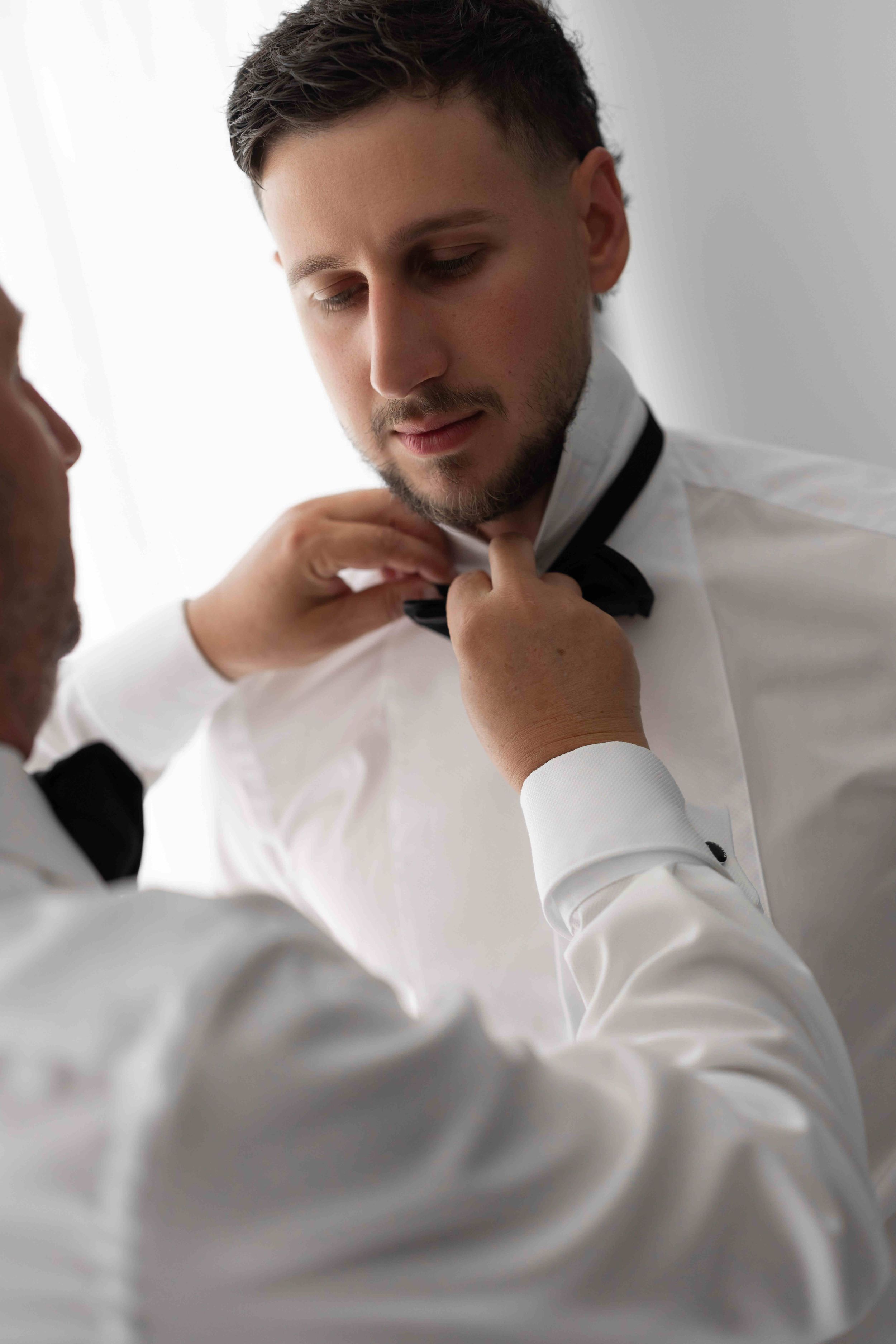 A man in a white tuxedo shirt gets his black bow tie adjusted by another person in a formal setting.