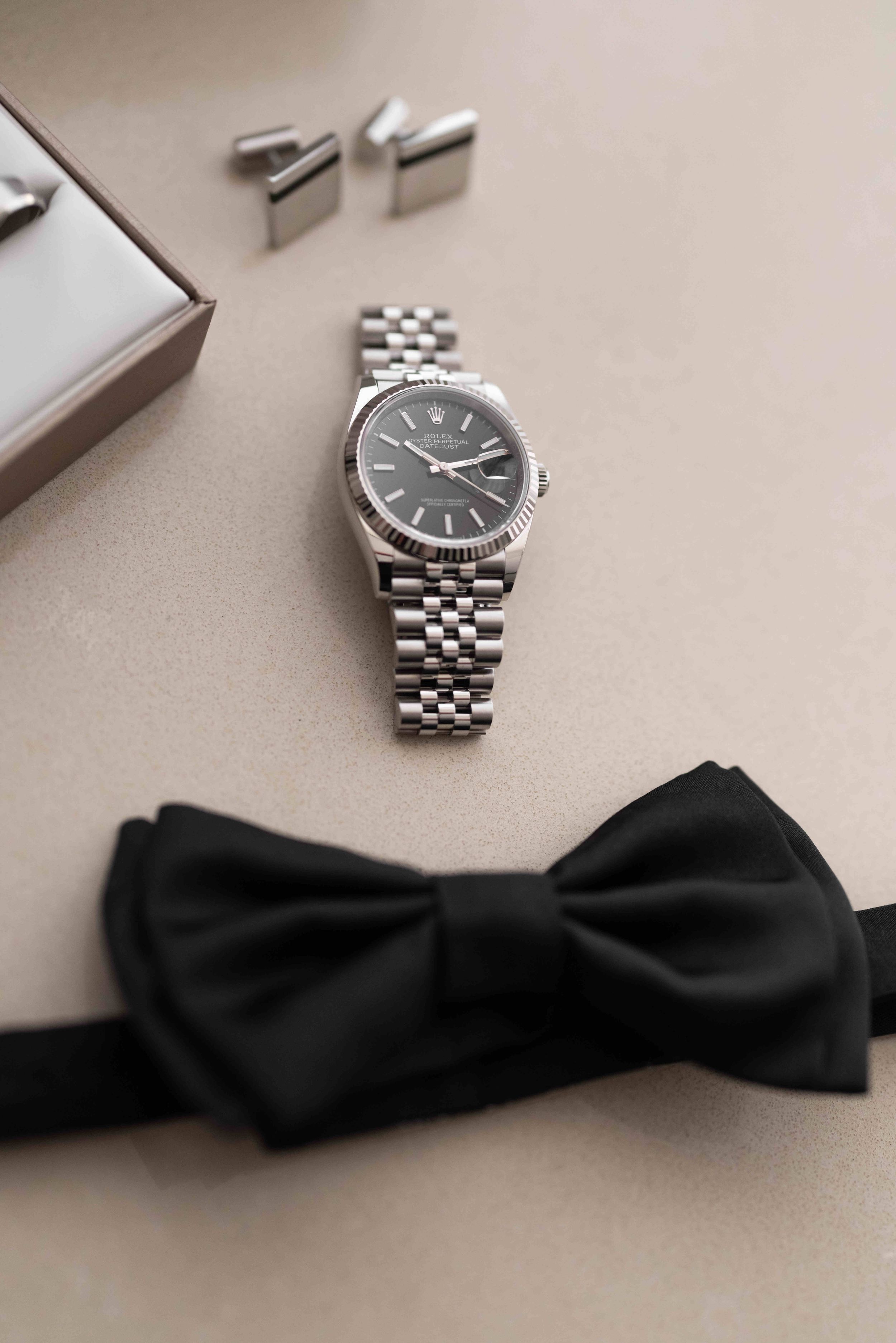 Silver Rolex Datejust watch with black dial beside a black bow tie and silver cufflinks on a beige surface.