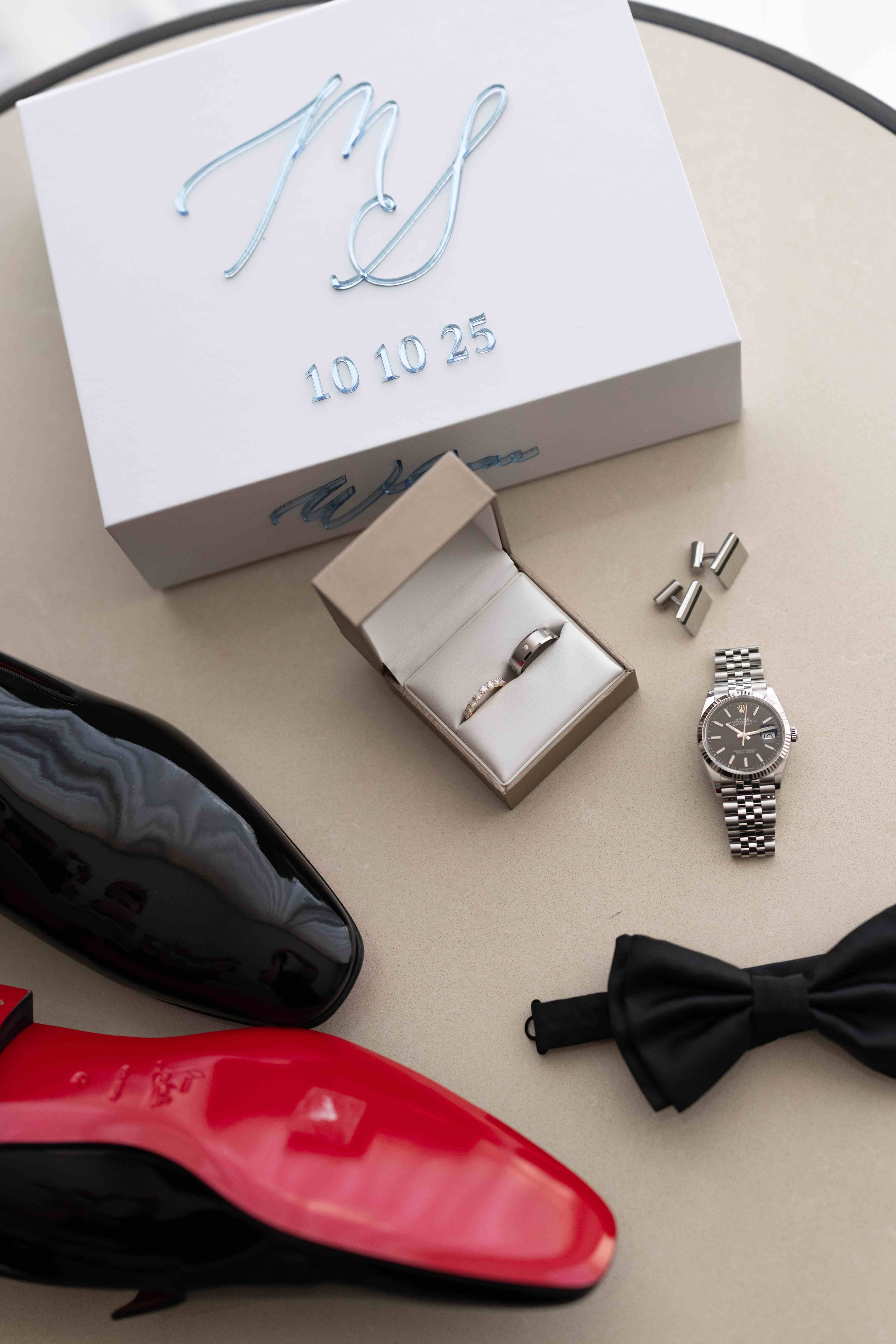 Groom wedding flat lay with black patent shoes, ring box, Rolex watch, cufflinks, bow tie, and monogrammed gift box.