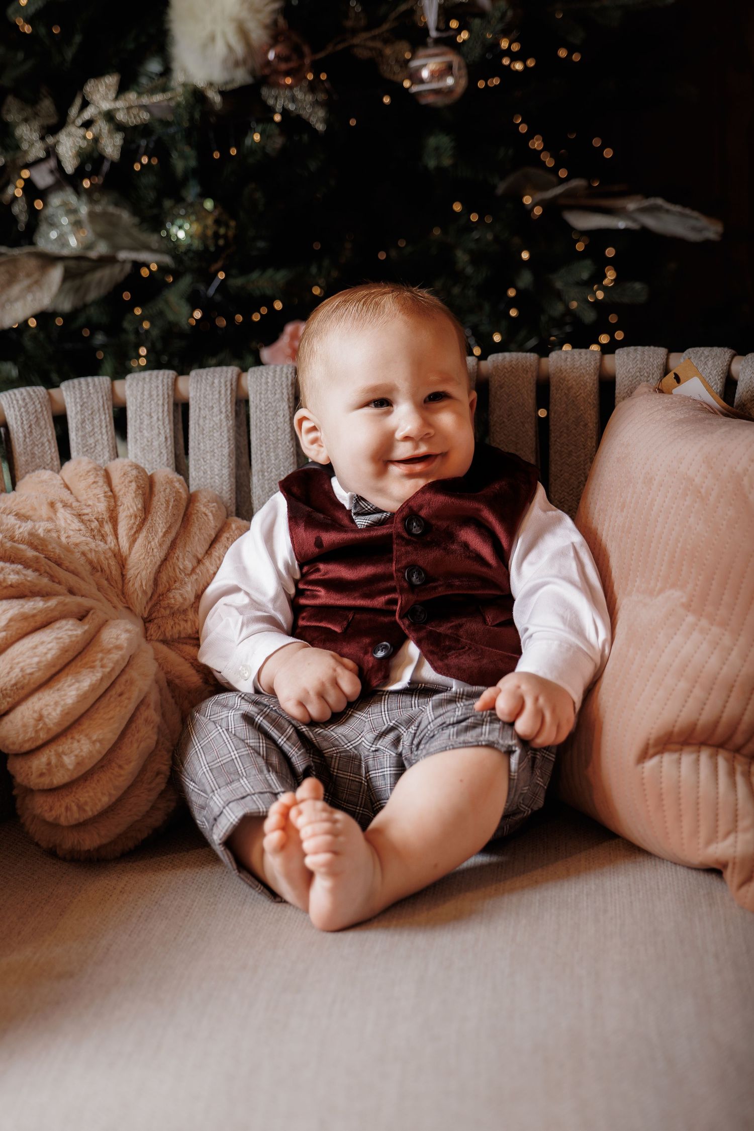 Smiling baby boy in burgundy vest and plaid shorts sits on couch beside Christmas tree with festive decorations.