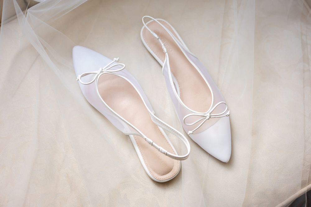 White satin pointed-toe slingback flats with delicate bow detail, perfect bridal wedding shoes on soft veil.