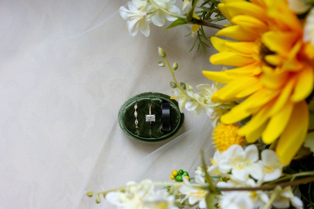 Green velvet ring box with diamond rings beside yellow flowers and white blooms on marble surface.