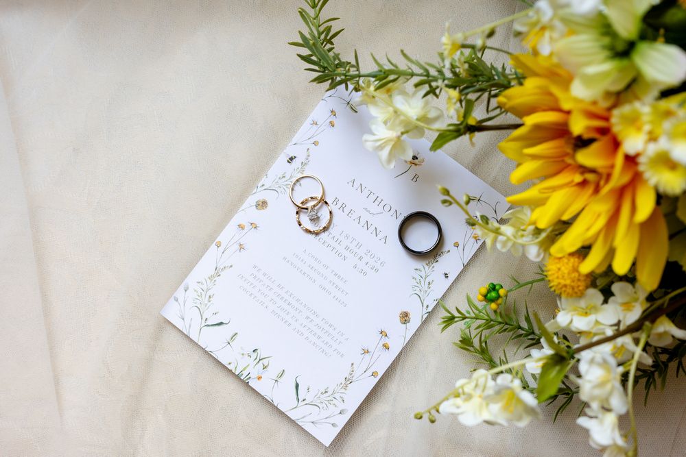 Wedding rings and bands resting on elegant floral invitation beside yellow sunflowers and white blooms.