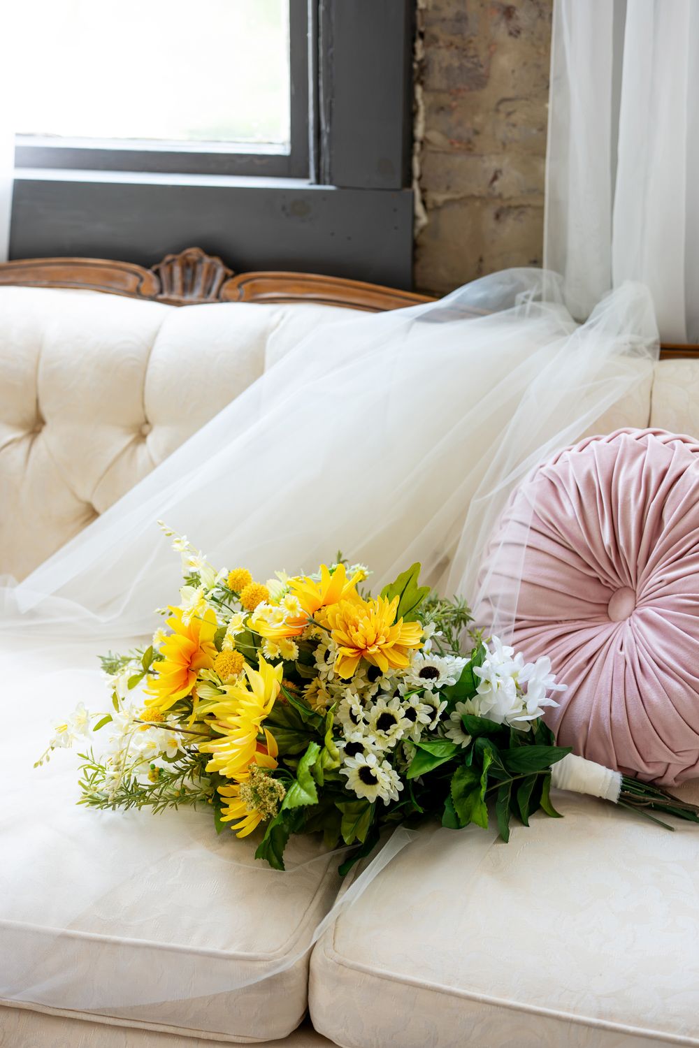 Yellow sunflower bridal bouquet resting on a white tufted sofa with a pink velvet pillow and wedding veil near a window.