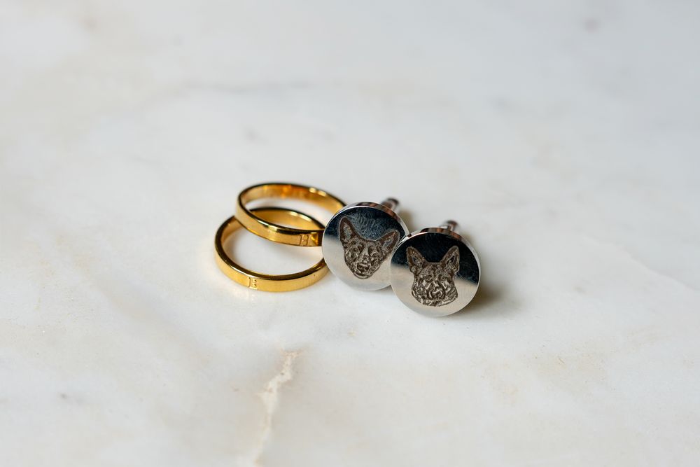 Gold wedding bands and silver French bulldog cufflinks displayed on a white marble surface.