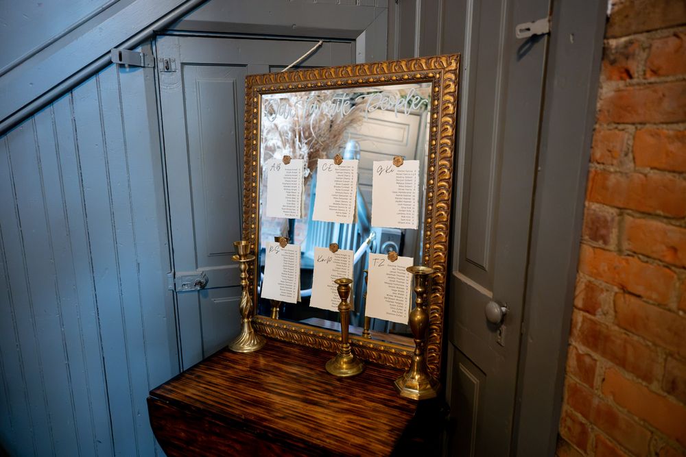 Gold ornate mirror with wedding seating chart cards displayed on a wooden table with brass candlesticks.