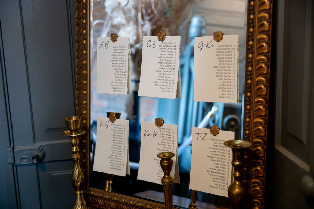 Gold-framed mirror displaying elegant wedding seating chart cards with brass candlesticks on a blue wall.